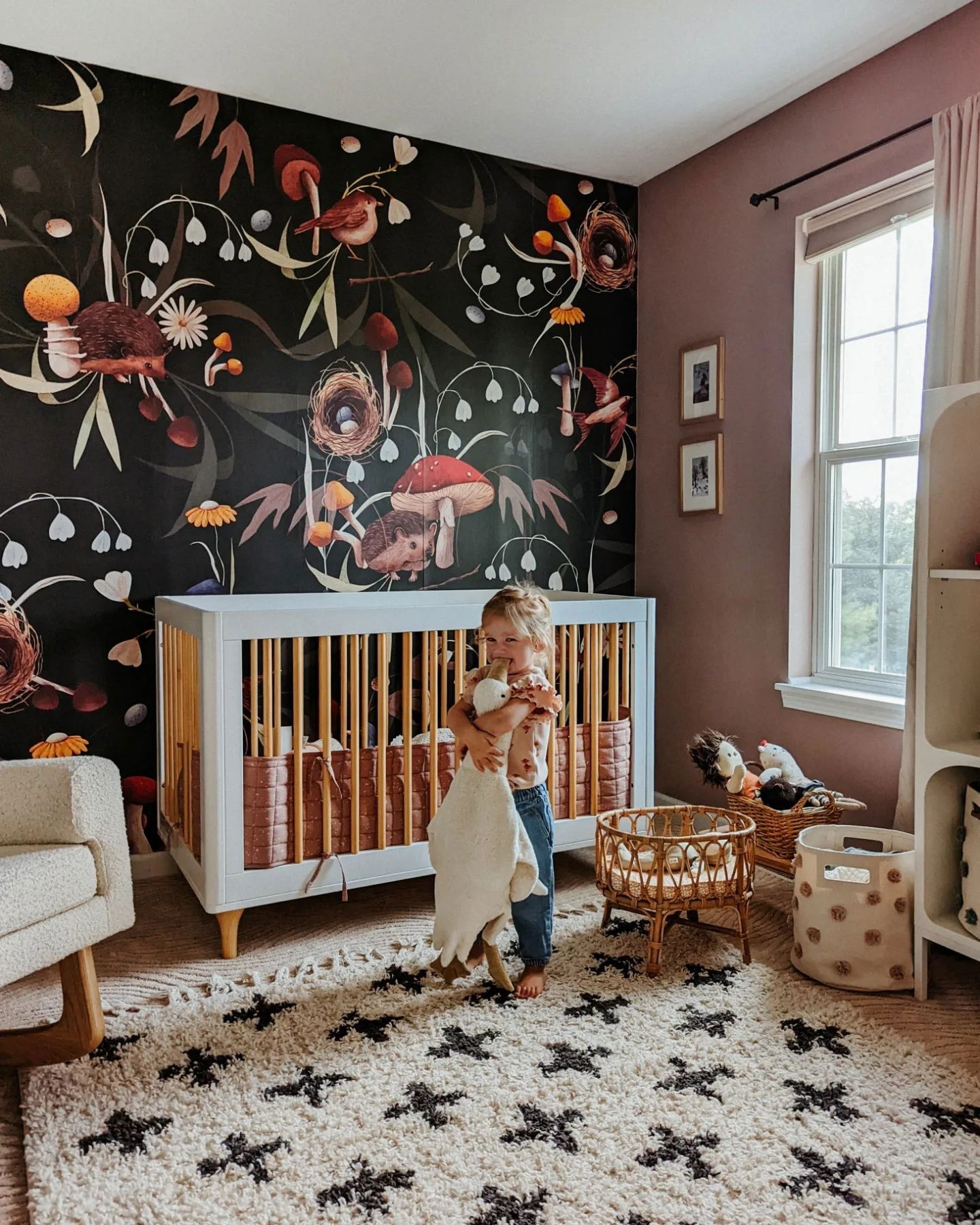 Kind umarmt ein Plüschtier in einem Kinderzimmer mit einer floralen Tapete, einem weißen Kinderbett und einem gemusterten Teppich.