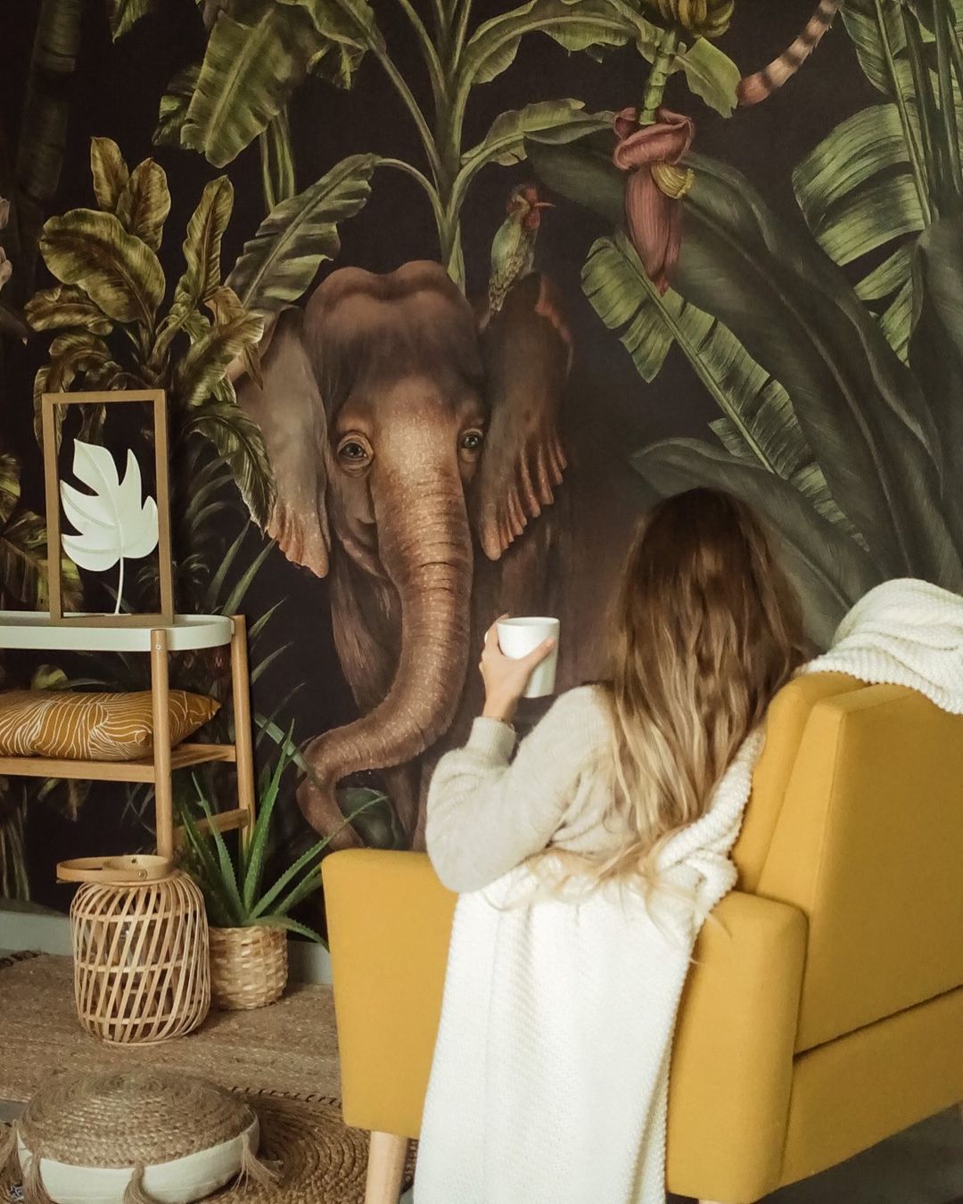 Woman with mug in yellow chair, blanket, and elephant jungle wallpaper.