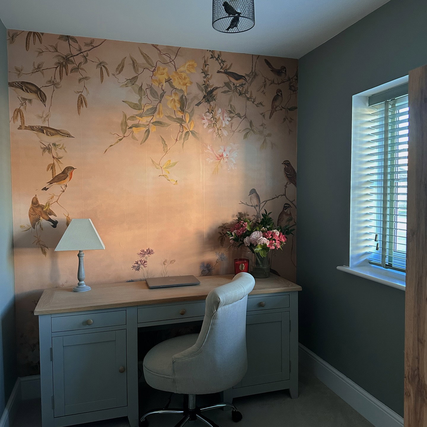 Een thuiskantoor met een lichtgrijs bureau en een gestoffeerde stoel. Een muur met perzikkleurig behang met vogels, gele en roze bloemen. Een lamp, laptop en bloemen staan op het bureau.