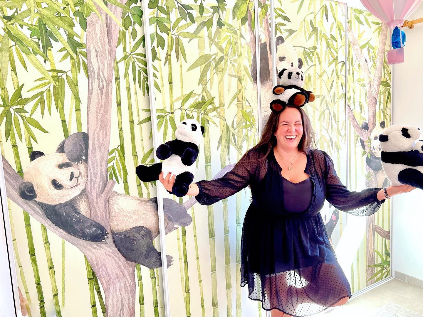 Een lachende vrouw in een zwarte jurk balanceert panda knuffels voor een muurschildering met panda thema.