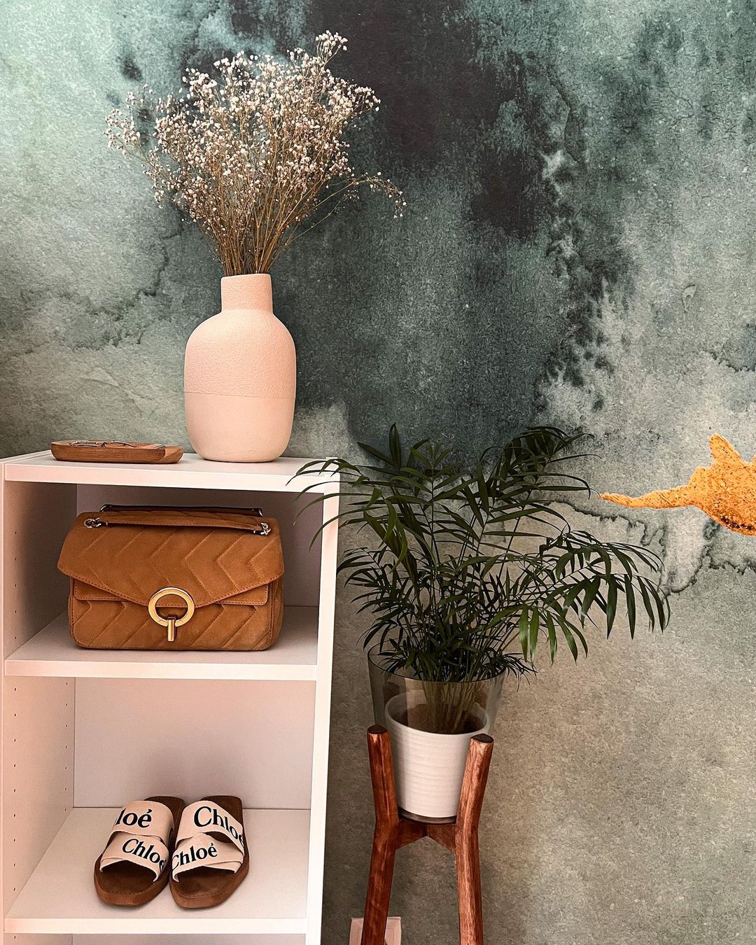 Witte planken met Chloé-slippers, bruine tas en vaas met bloemen. Potplant op houten standaard.