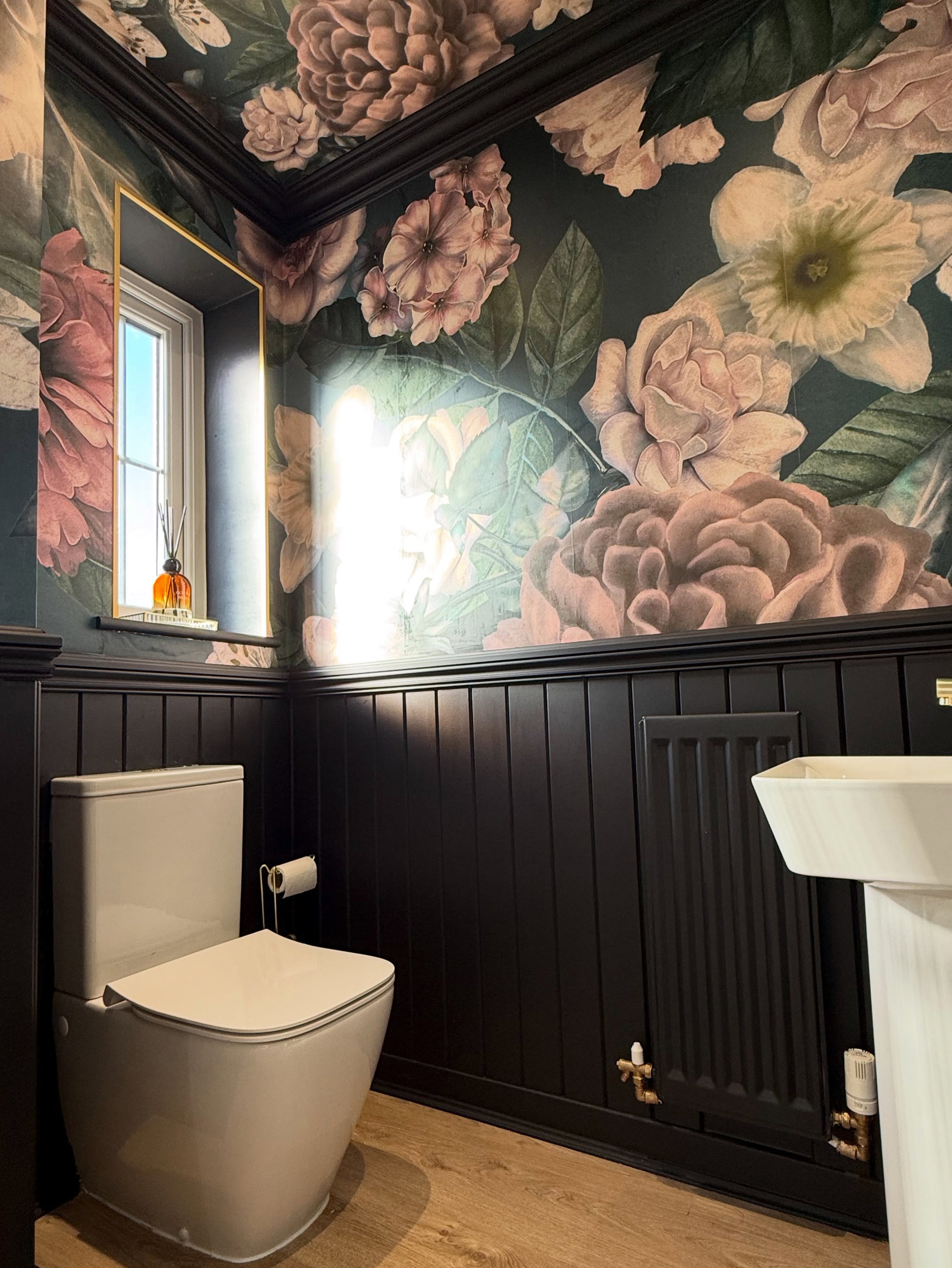 Et badeværelse med mørkt blomstret tapet med pink og hvide blomster over mørkt panelbeklædt brystning. Et lysfarvet toilet, et mørkt radiator og et delvist synligt hvidt piedestalvask er synlige. Et lille vindue med et guldfarvet spejl er til venstre.