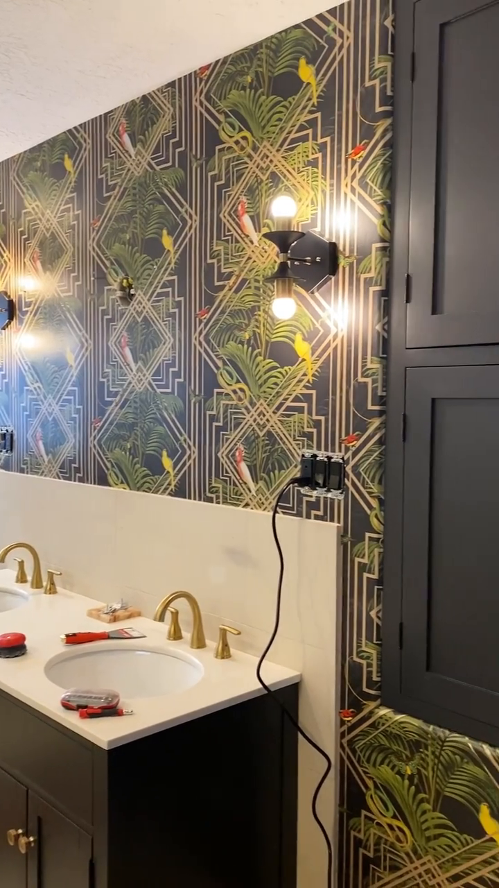 Bathroom with double sink, gold faucets, tropical wallpaper, and black cabinets.