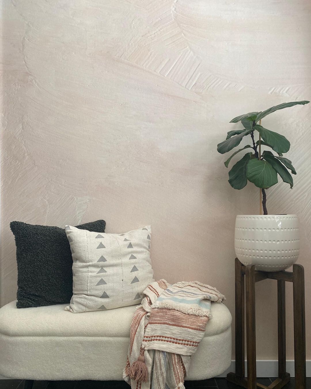 Interior with textured pink wall, bench with pillows and throw, and potted fiddle-leaf fig on a stand.