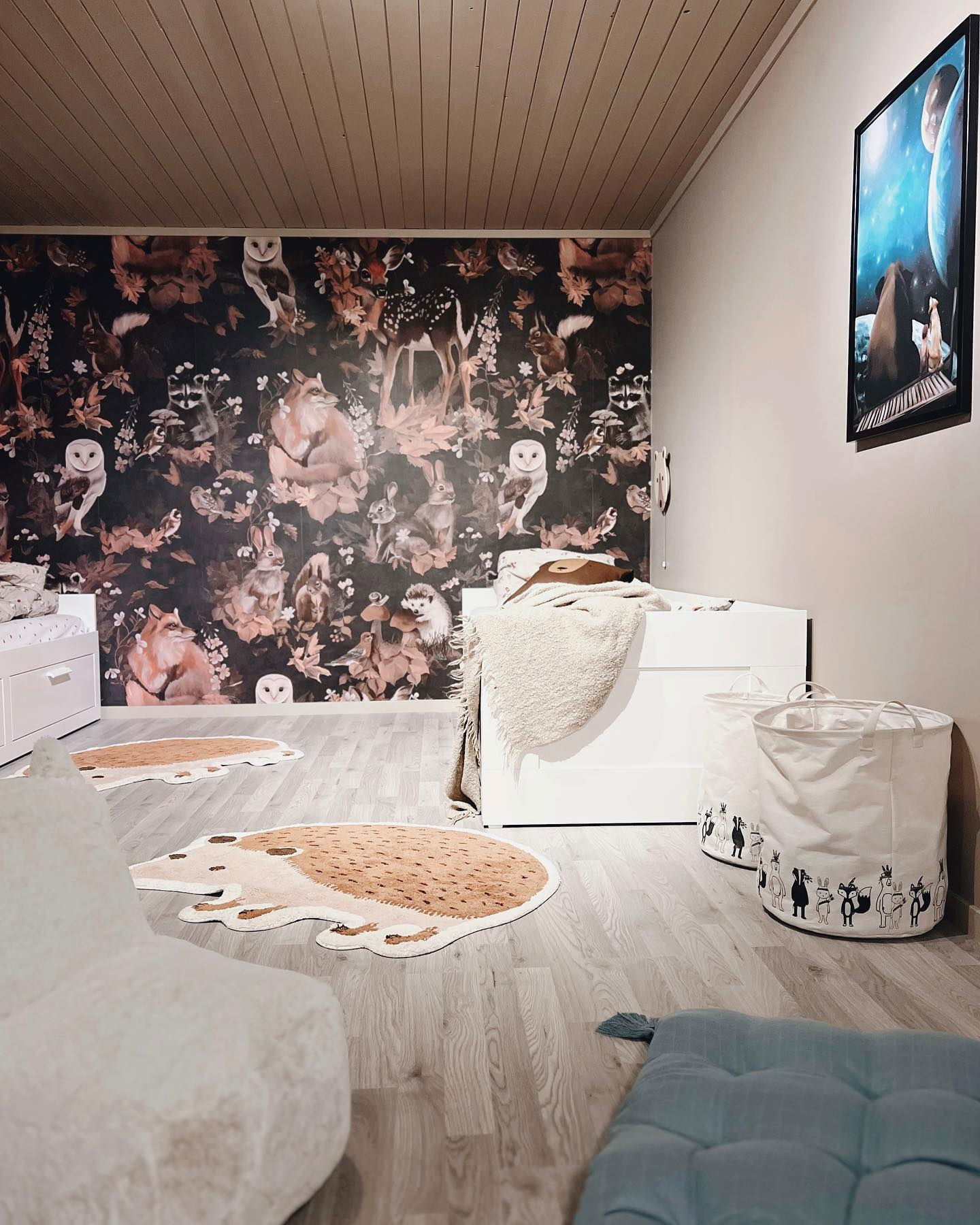 Chambre d'enfant avec papier peint sur le thème de la forêt, tapis hérisson, bacs de rangement blancs et une reproduction d'art spatial encadrée.