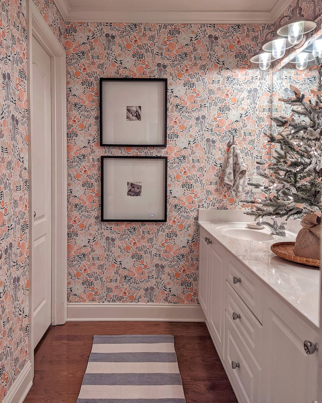 Badrum med blommig tapet, vitt tvättställ och inramade babyfoton.