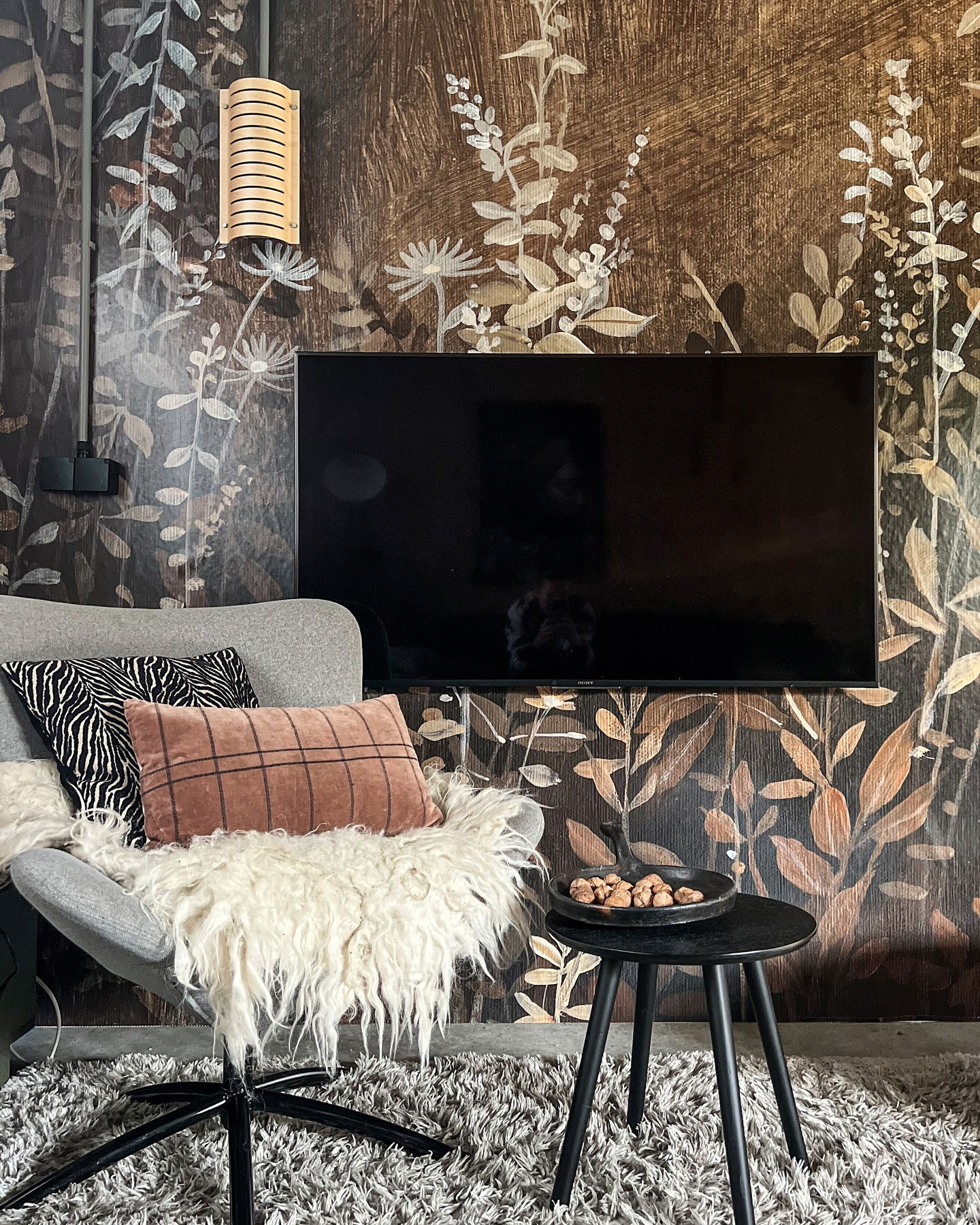 Living room with floral wallpaper, TV, gray chair with pillows and sheepskin, and small black table.
