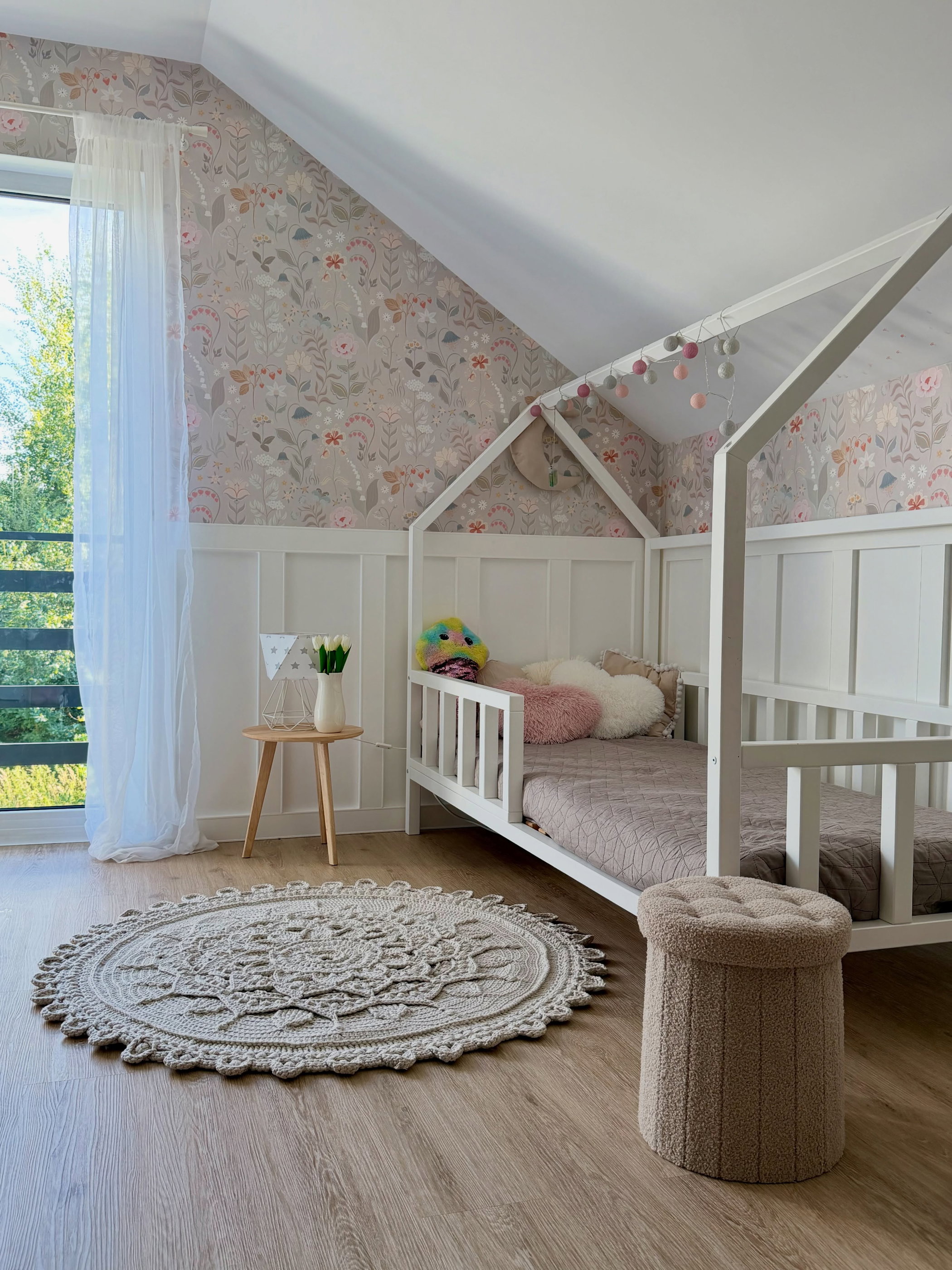 A child's room with a white house-shaped bed, beige floral wallpaper, white wainscoting, and a round crocheted rug on a wooden floor.