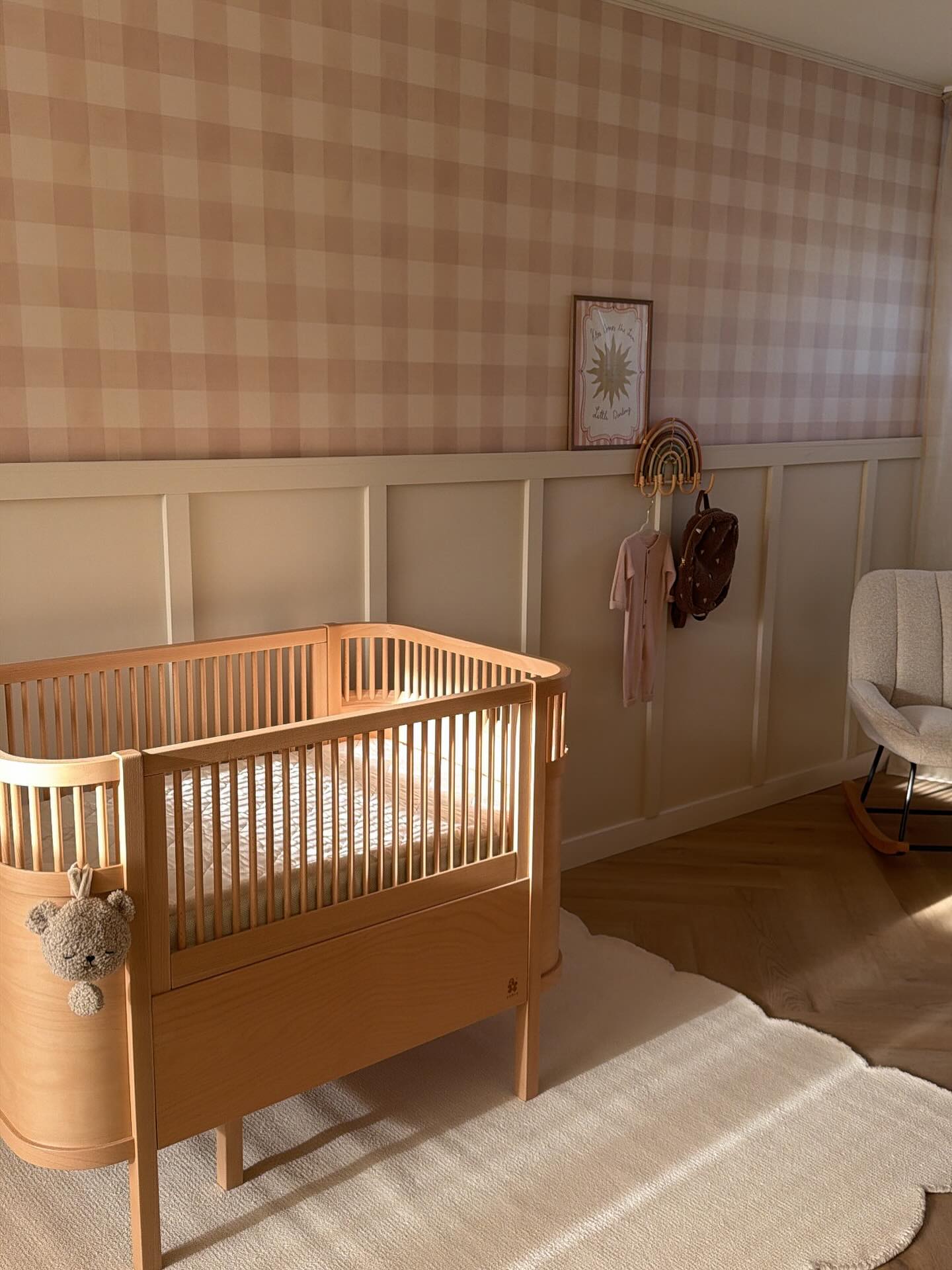 Light wood crib with teddy bear decoration in a nursery with gingham wallpaper and a rocking chair.