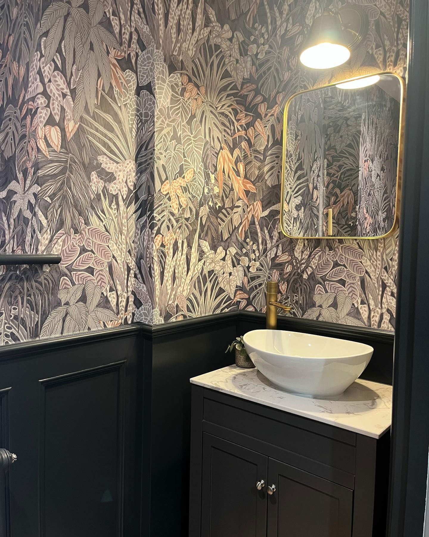 Bagno con carta da parati tropicale, boiserie nera e lavabo a catino bianco su mobile nero.