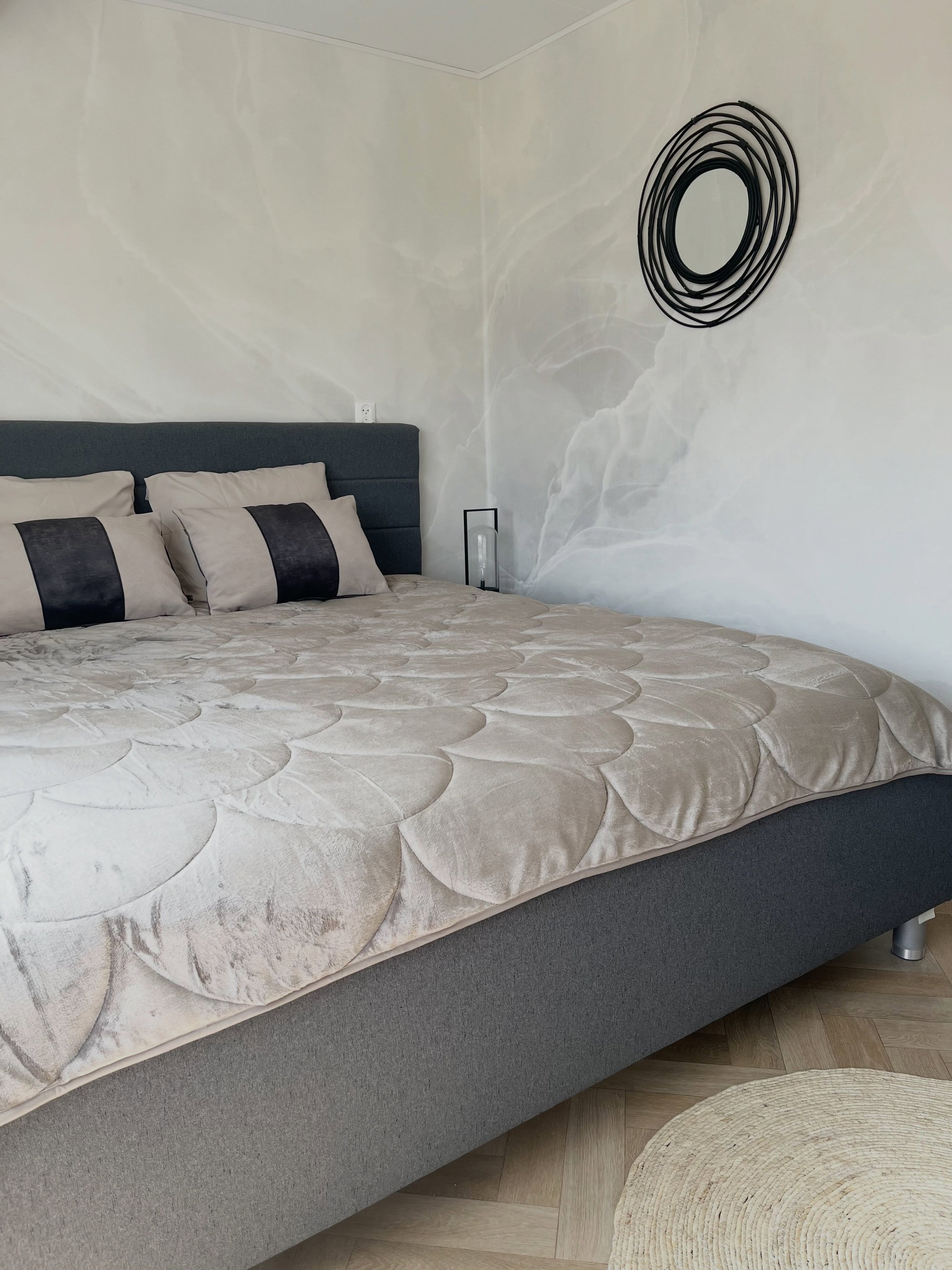 Modern bedroom with gray upholstered bed, tan quilted duvet, decorative pillows, and abstract wall art.