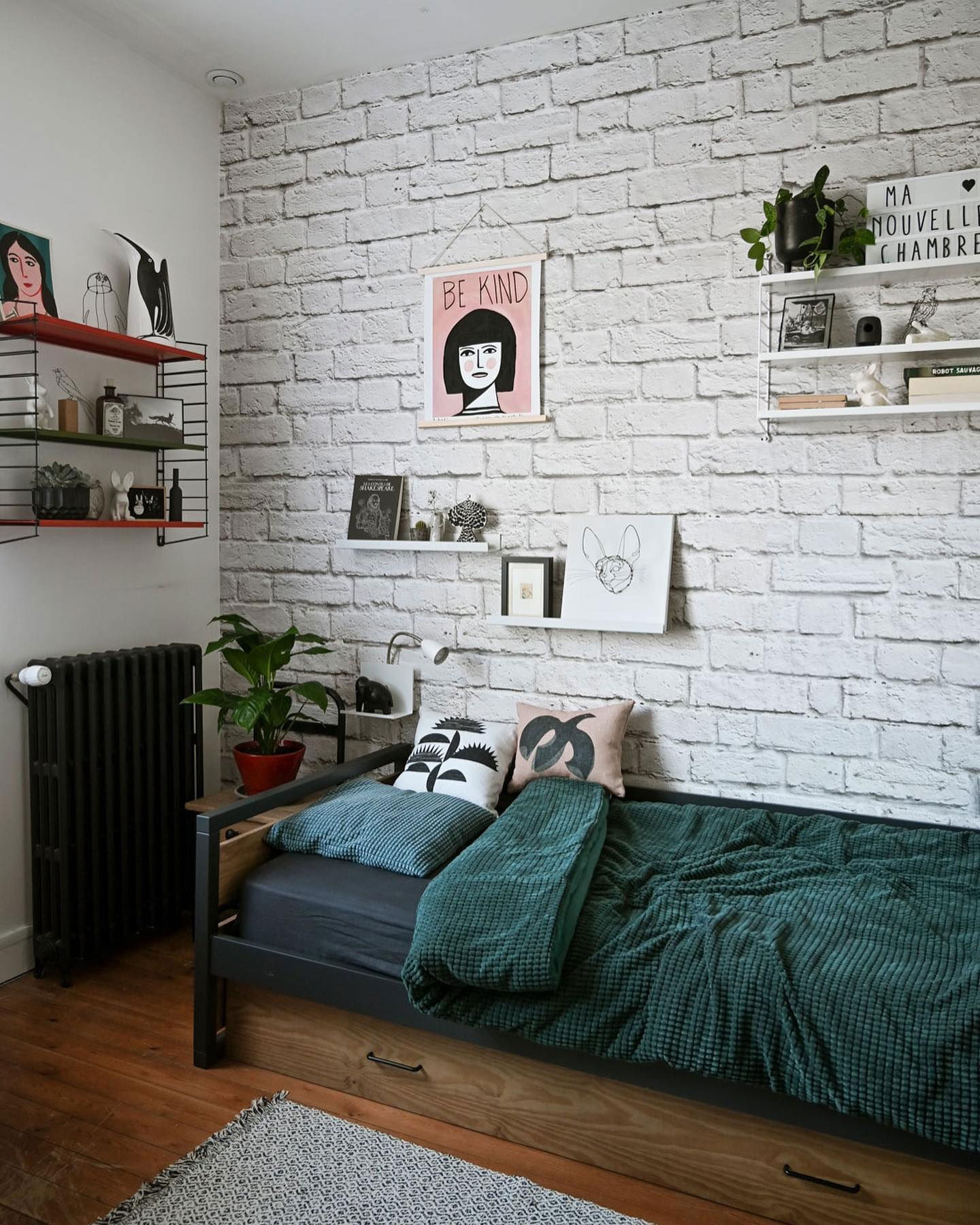 Bedroom with bed against white brick wall, decorated with shelves and art.