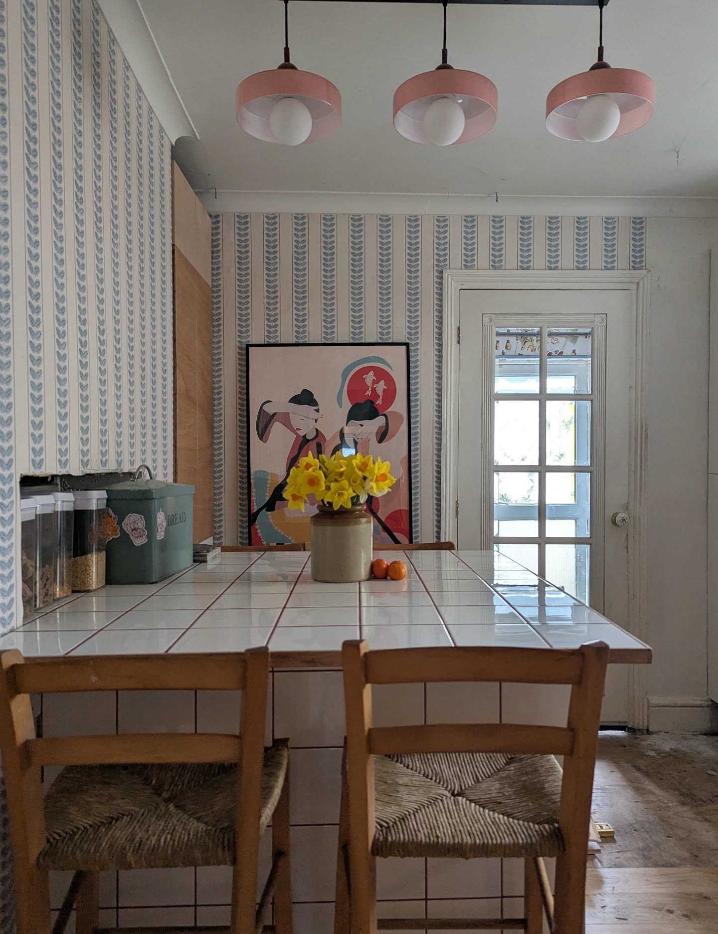 Kitchen island with white tile top, two wooden chairs, daffodils, and artwork.