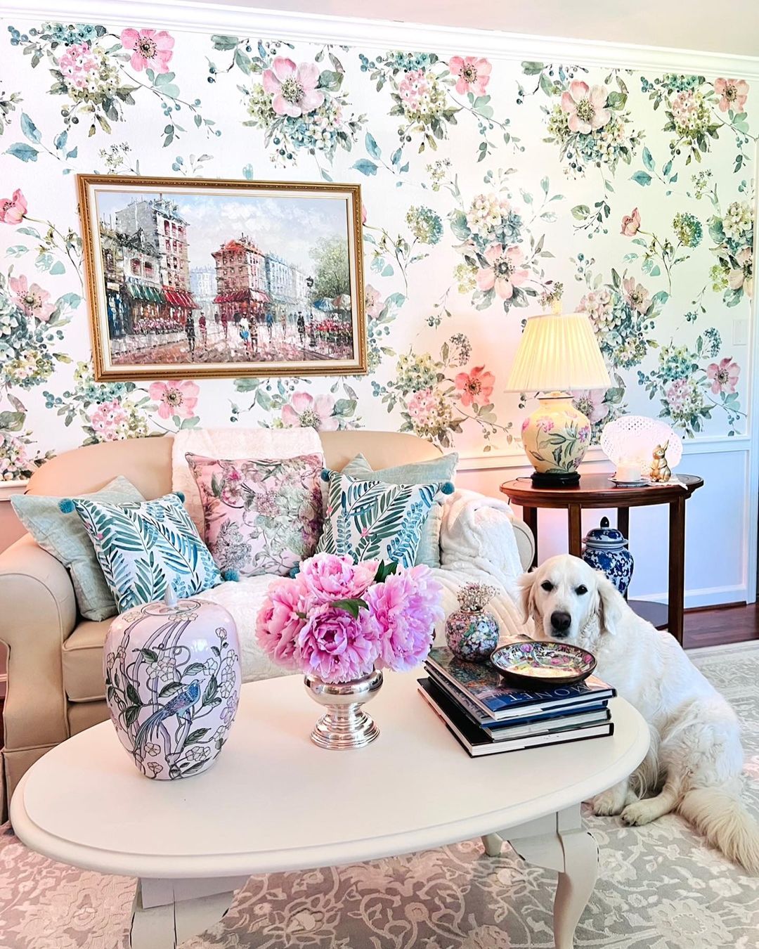 Wohnzimmer mit floraler Tapete, Gemälde und weißem Sofa. Ein Golden Retriever sitzt an einem weißen Couchtisch mit Büchern und rosa Pfingstrosen.