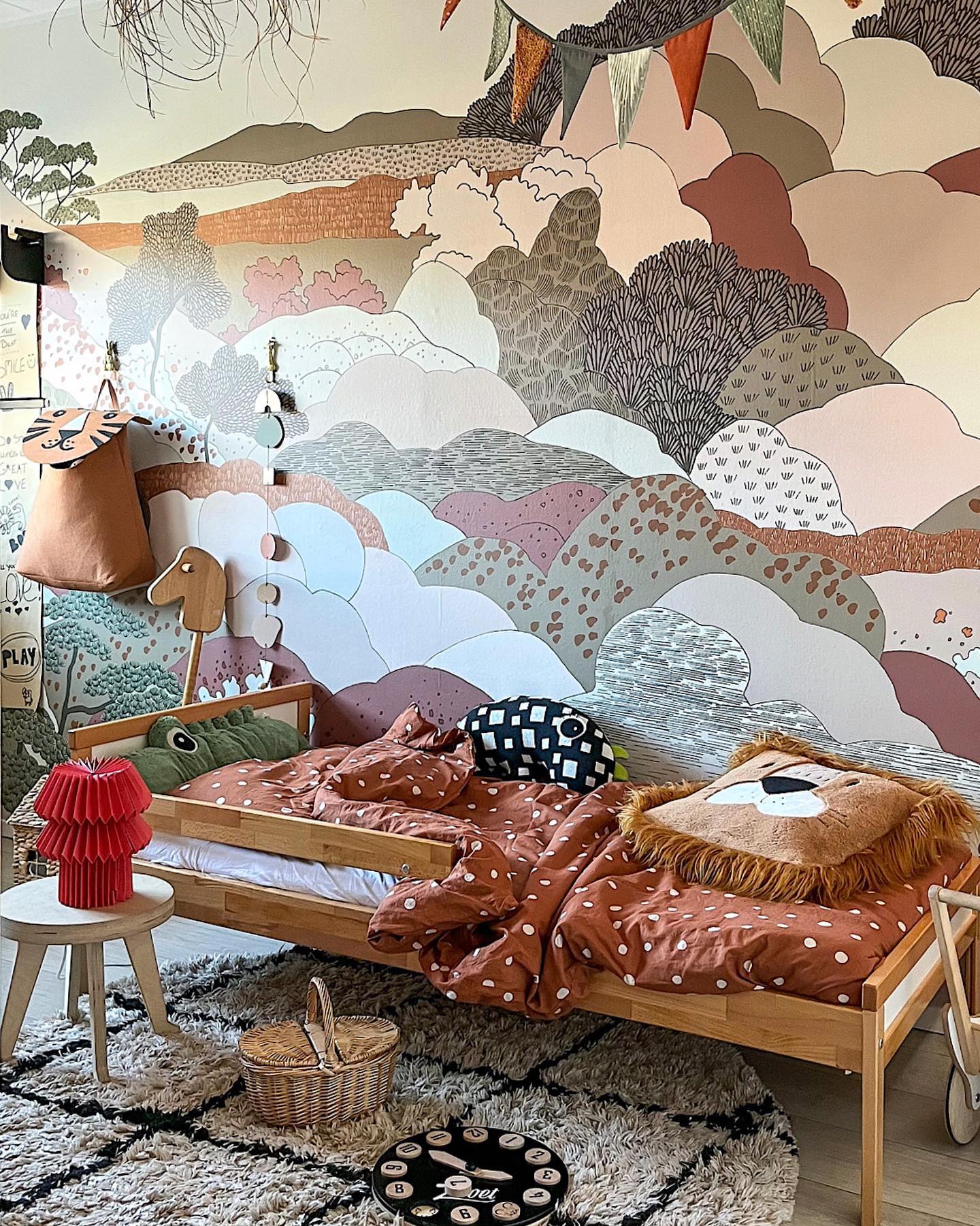 A child's bedroom features a wooden bed with brown polka dot bedding, a lion cushion, and a green crocodile pillow. The wall has a colorful mural of rolling hills and trees.