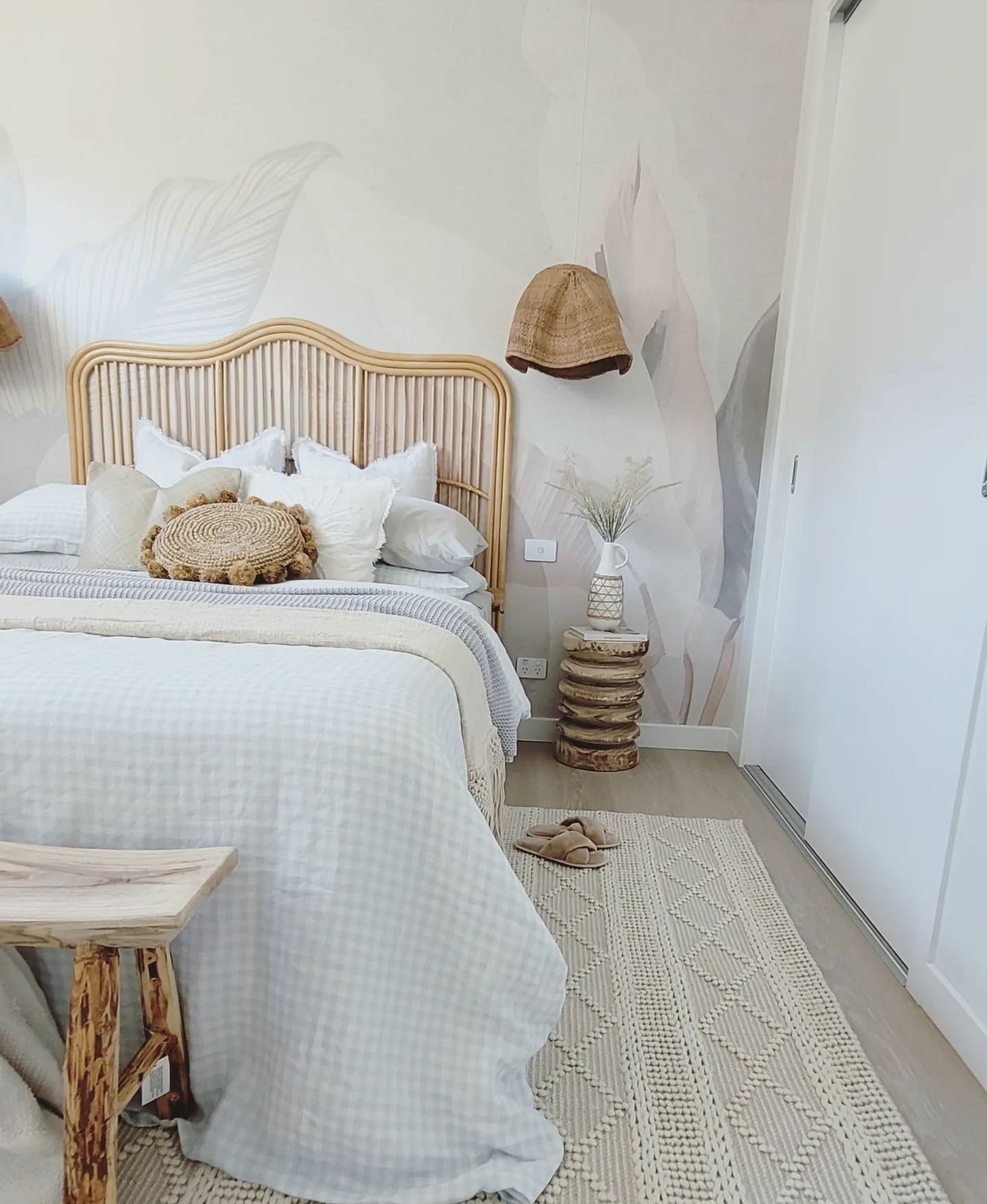Bedroom with rattan headboard, white bedding, and leaf-patterned wallpaper. A woven rug and wooden stool add texture.