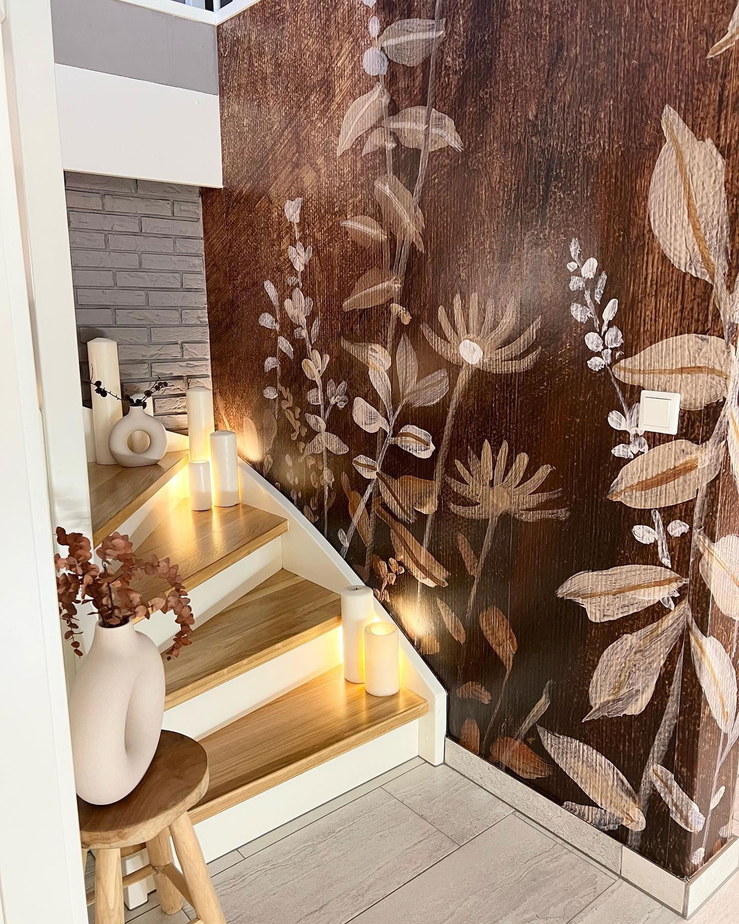 Staircase with wood steps, white risers, and floral wallpaper. Candles illuminate the steps. A vase sits on a wooden stool.