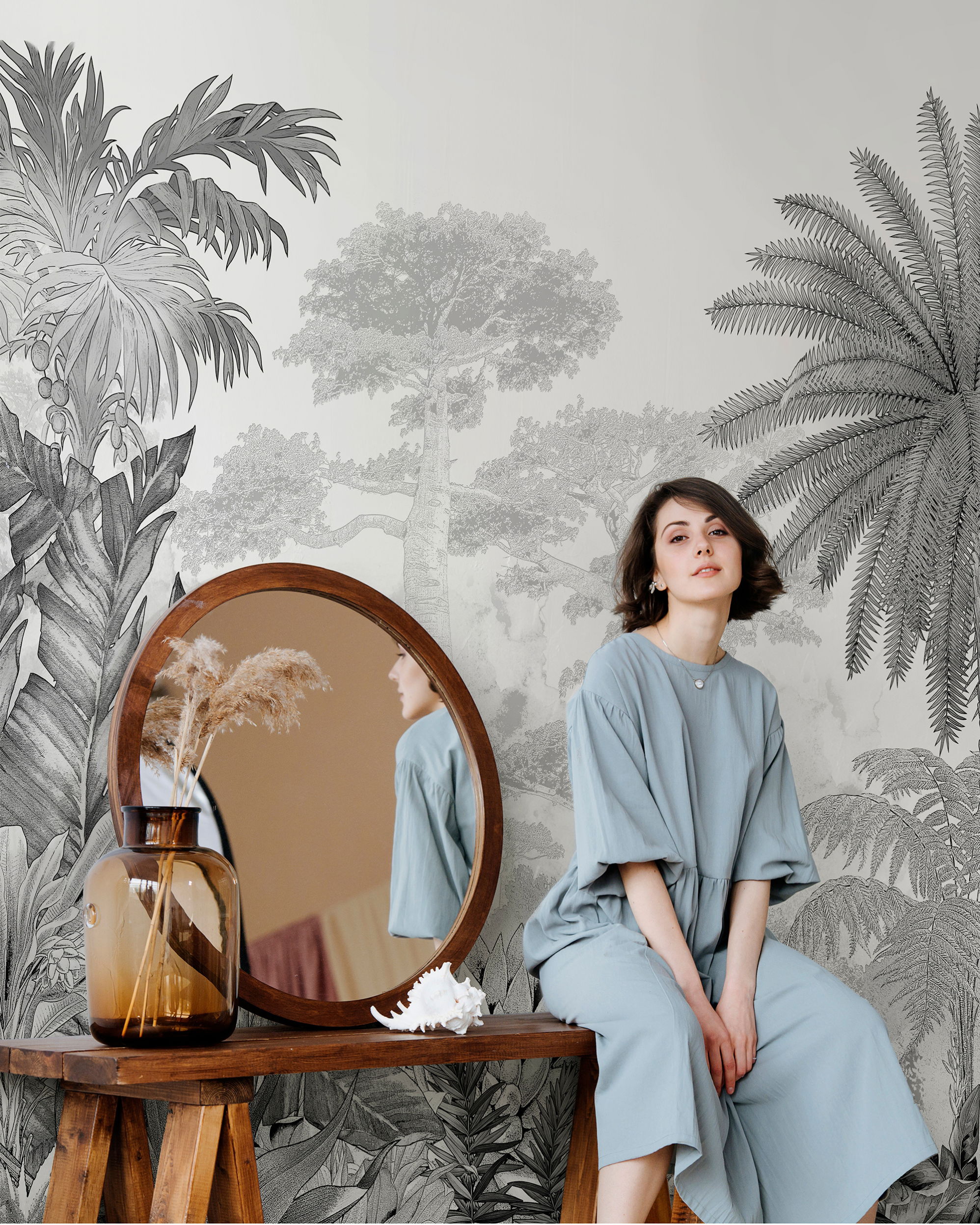 A woman in a blue outfit sits near a mirror and tropical wallpaper.