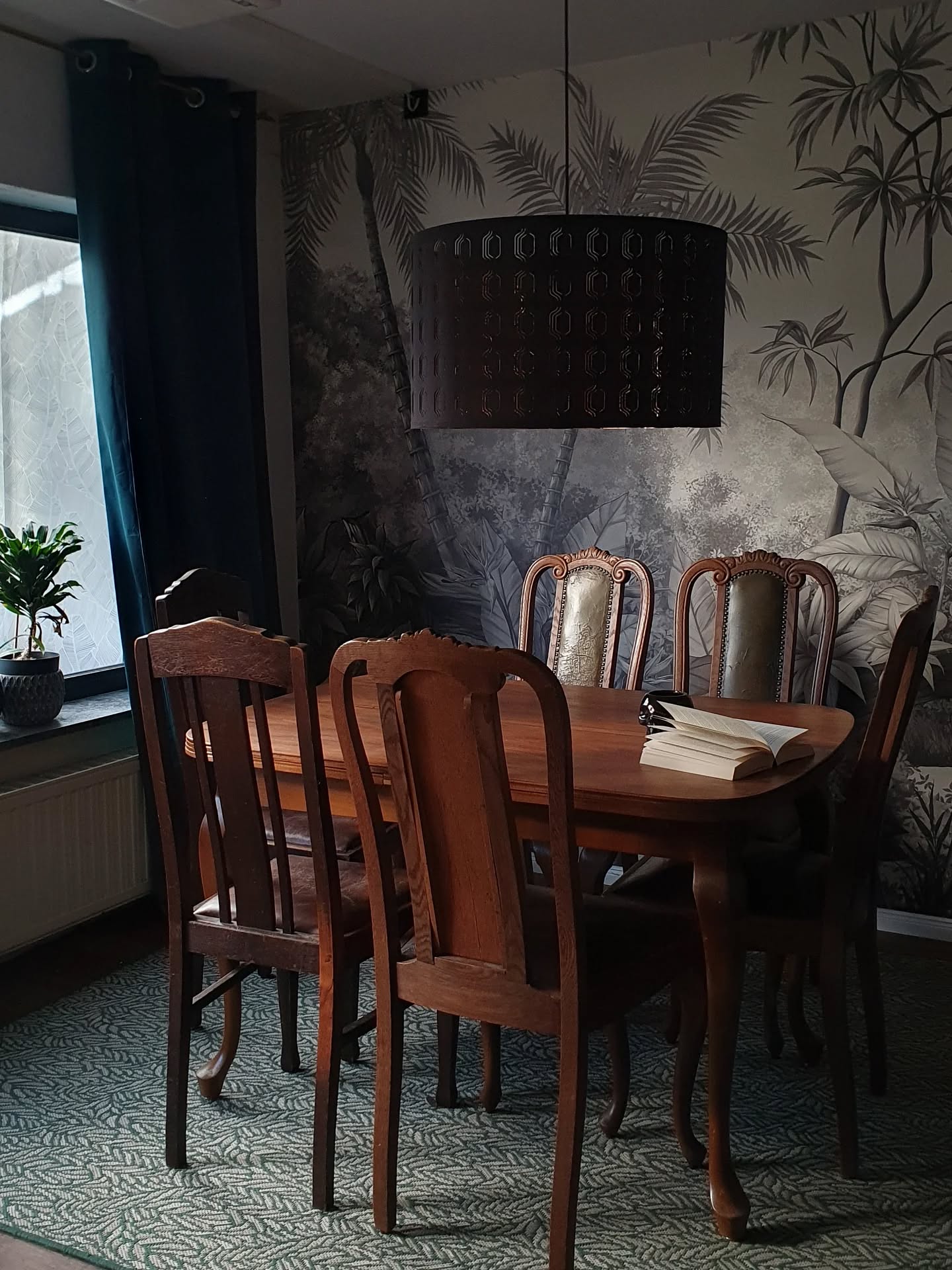En spisestue med et trebord og stoler, en mørk trommelykt, svart og hvit jungeltapet og et grønt mønstret teppe.