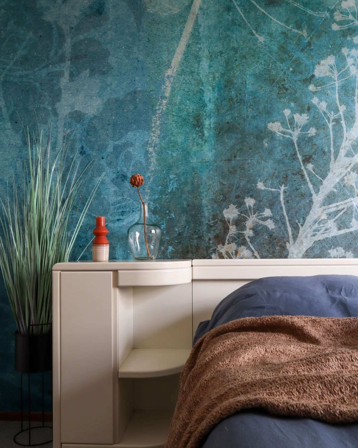 Bedroom interior with blue floral wallpaper, white headboard, and brown throw blanket.