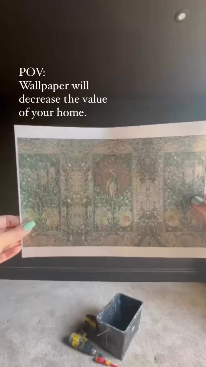 Close-up of hands holding floral wallpaper sample, with tools and paint can below.