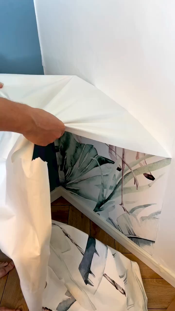 Hand peeling back white paper to reveal tropical leaf wallpaper on a wall.