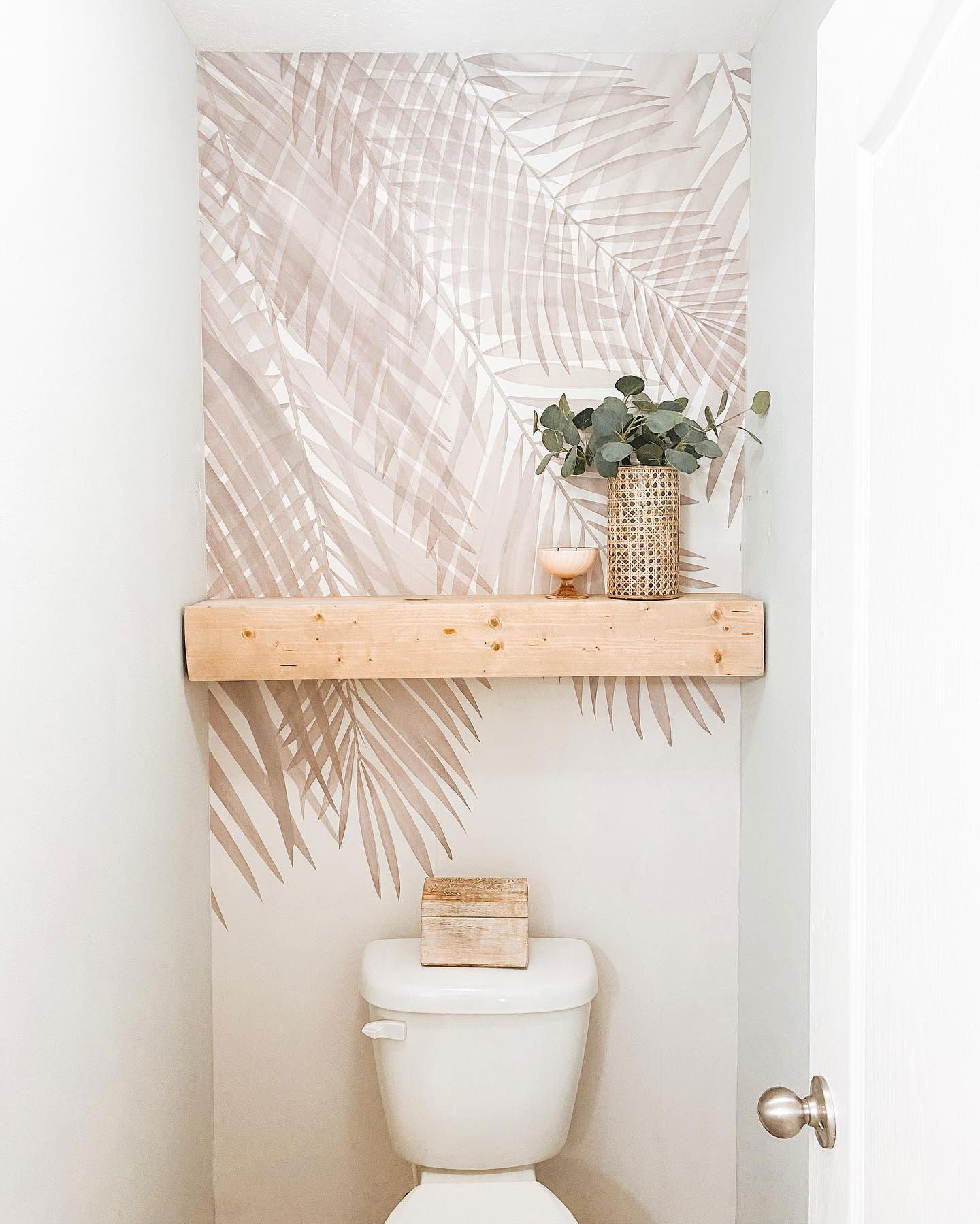 Baño pequeño con papel tapiz de hojas de palma, estante de madera e inodoro blanco. Un jarrón tejido con vegetación se encuentra en el estante.