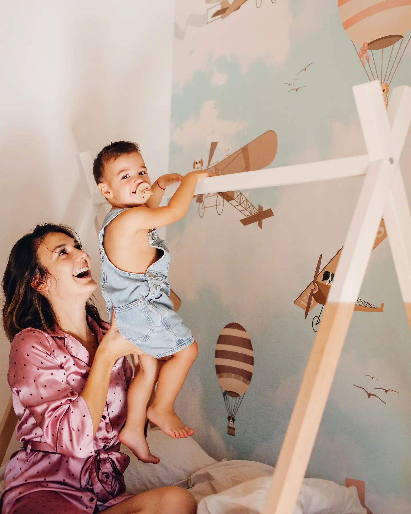 A mother holds up her toddler in a bedroom with airplane wallpaper. The child wears denim overalls and has a pacifier.
