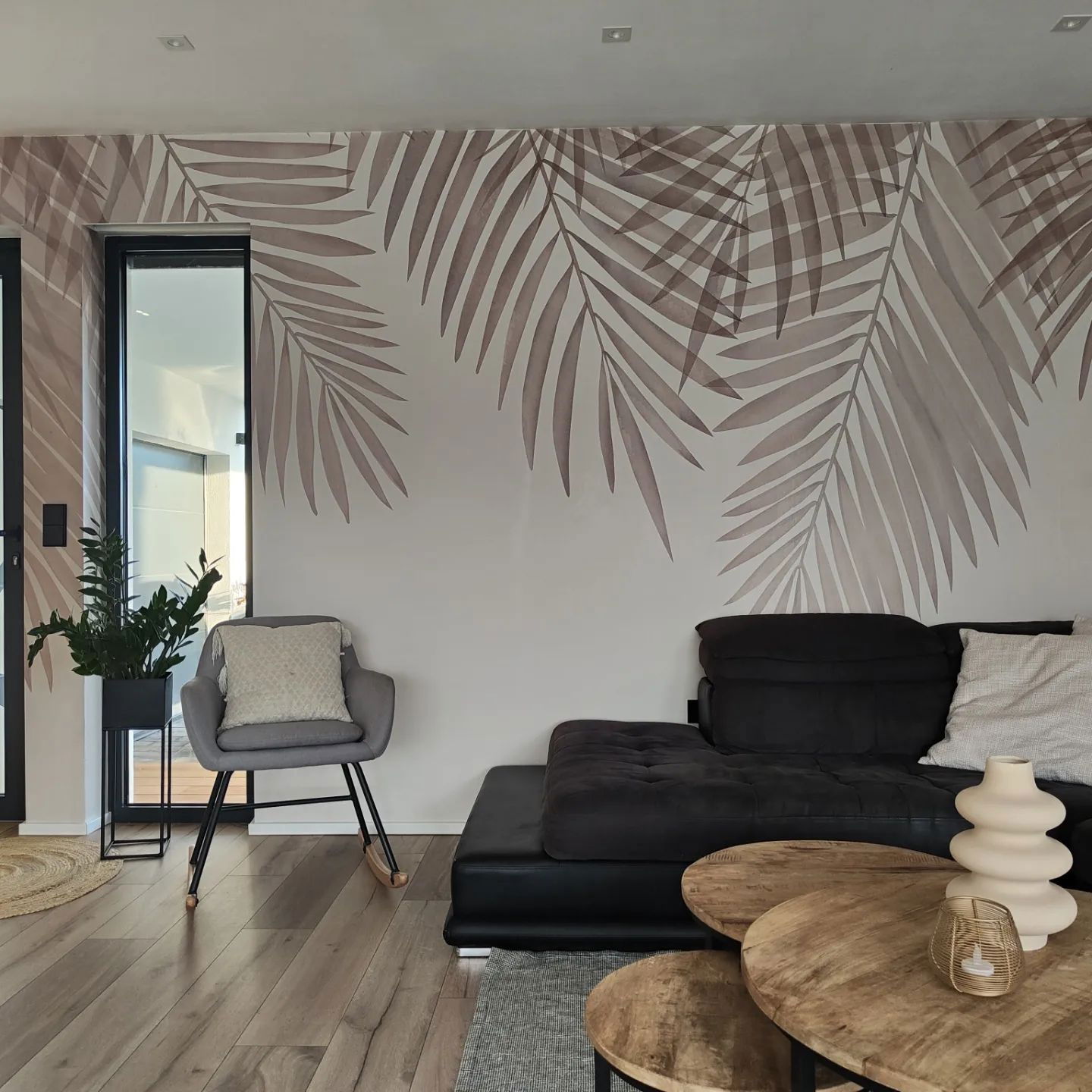 Sala de estar con papel tapiz de hojas de palma, mecedora gris y sofá modular negro.