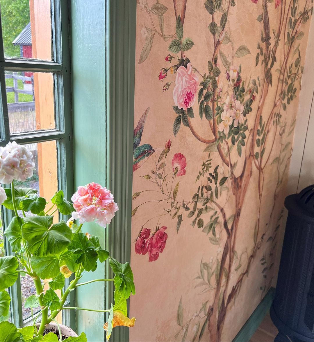 Indvendig udsigt med blomstret tapet, pelargonier i et vindue og et glimt af en rød bygning udenfor.