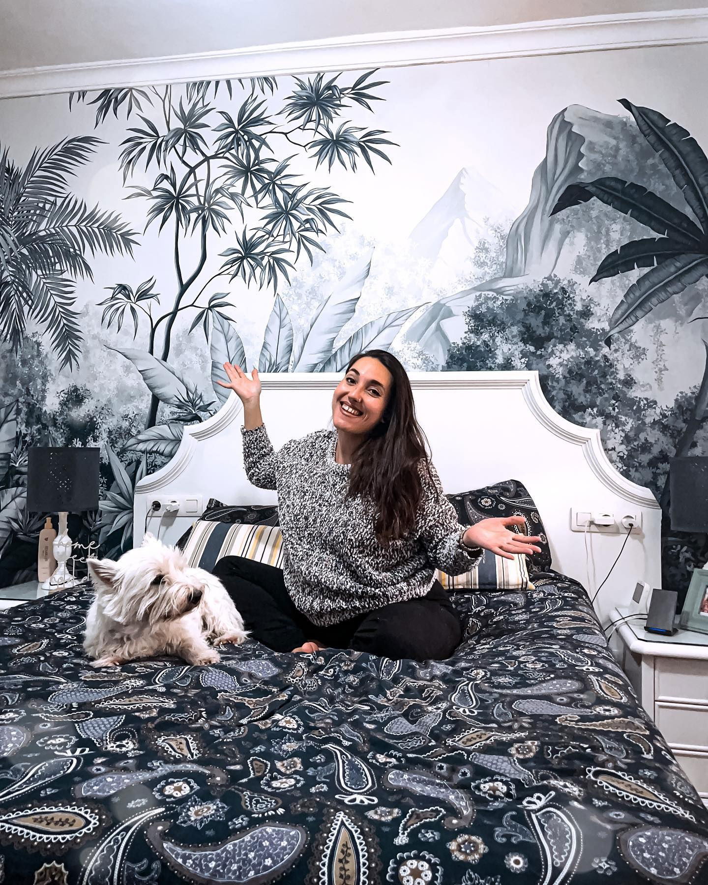 A smiling woman sits cross-legged on a paisley bedspread with a Westie dog. A black and white tropical mural is behind the bed.