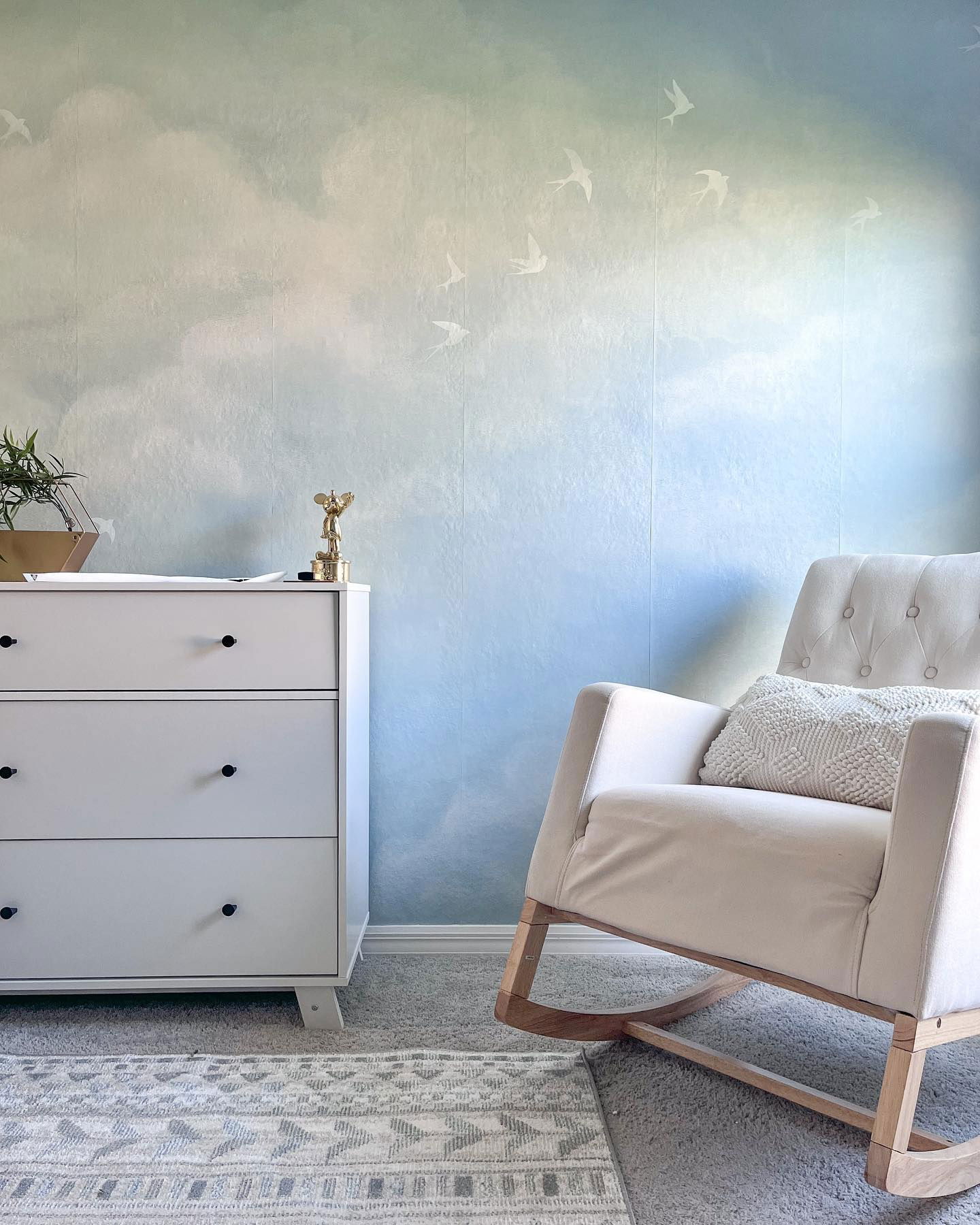 Nursery with white dresser, rocking chair, and cloud wallpaper with birds.