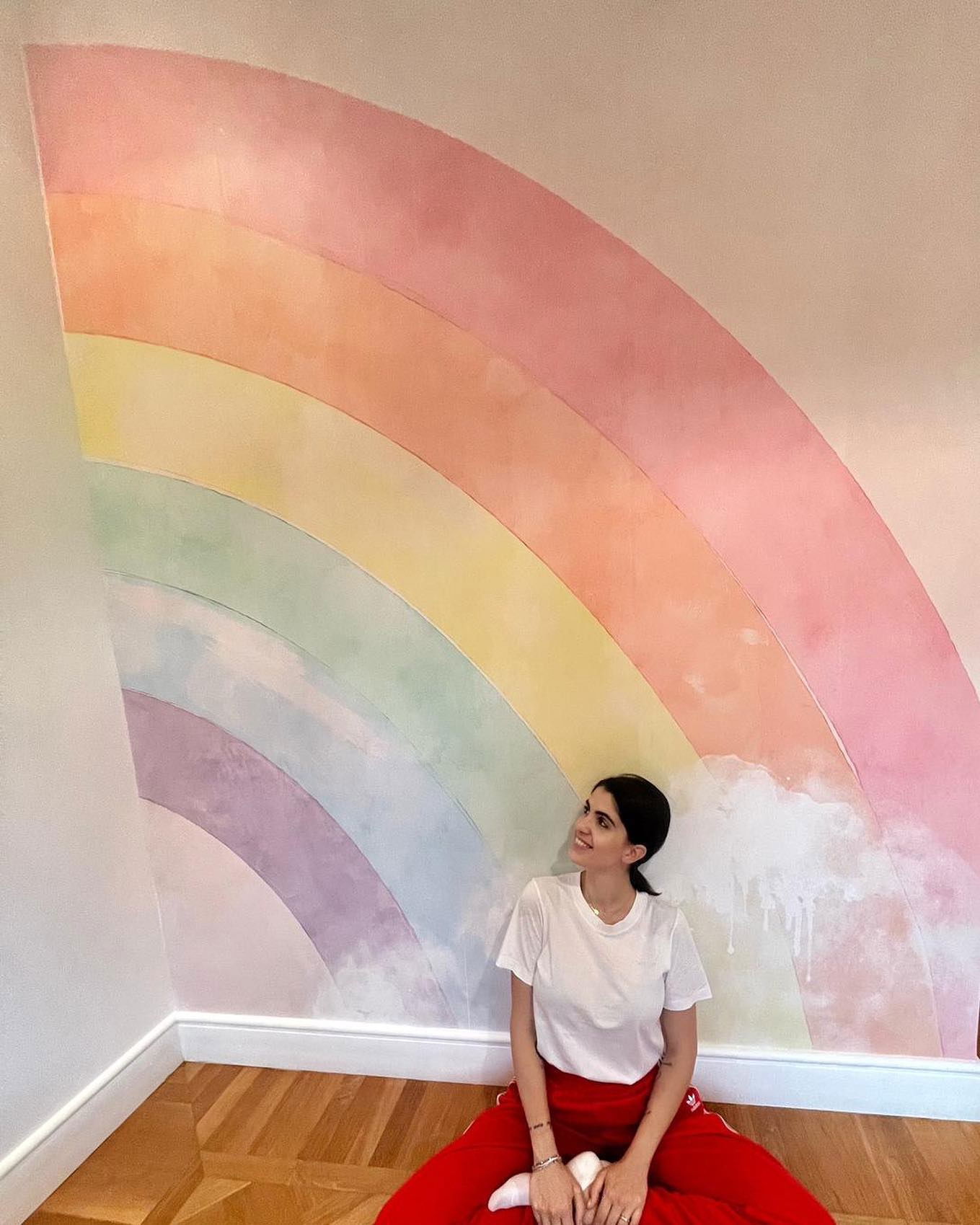 A woman sits in front of a pastel rainbow painted on a wall.