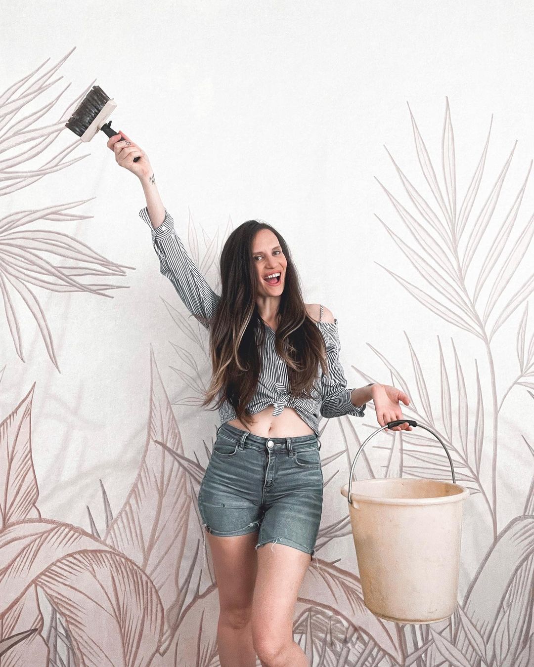 A woman holding a brush and bucket stands in front of a wall with a leaf pattern.