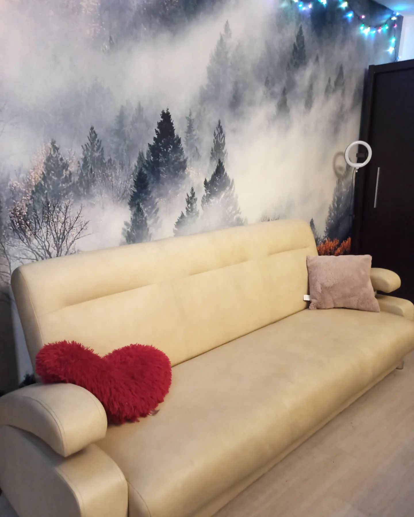 Beżowa sofa z poduszkami w kształcie serca i kwadratu na tle muralu z motywem lasu.