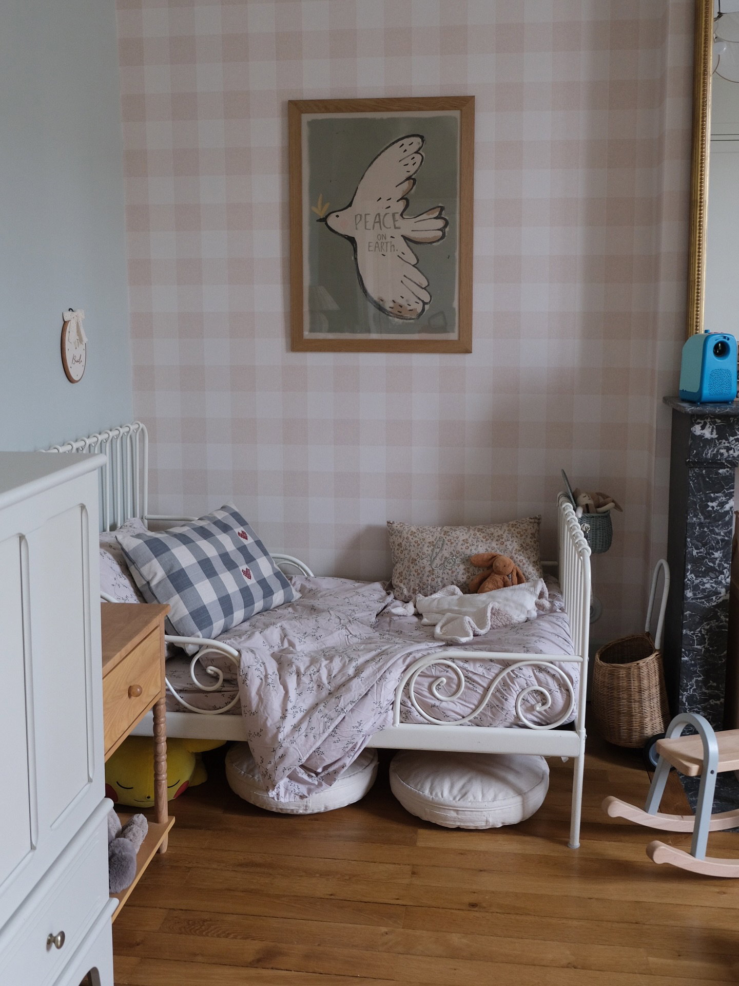 Et børneværelse med en hvid metalseng, lyserødt og hvidt ternet tapet og et indrammet tryk af en hvid due med "PEACE" over sengen.