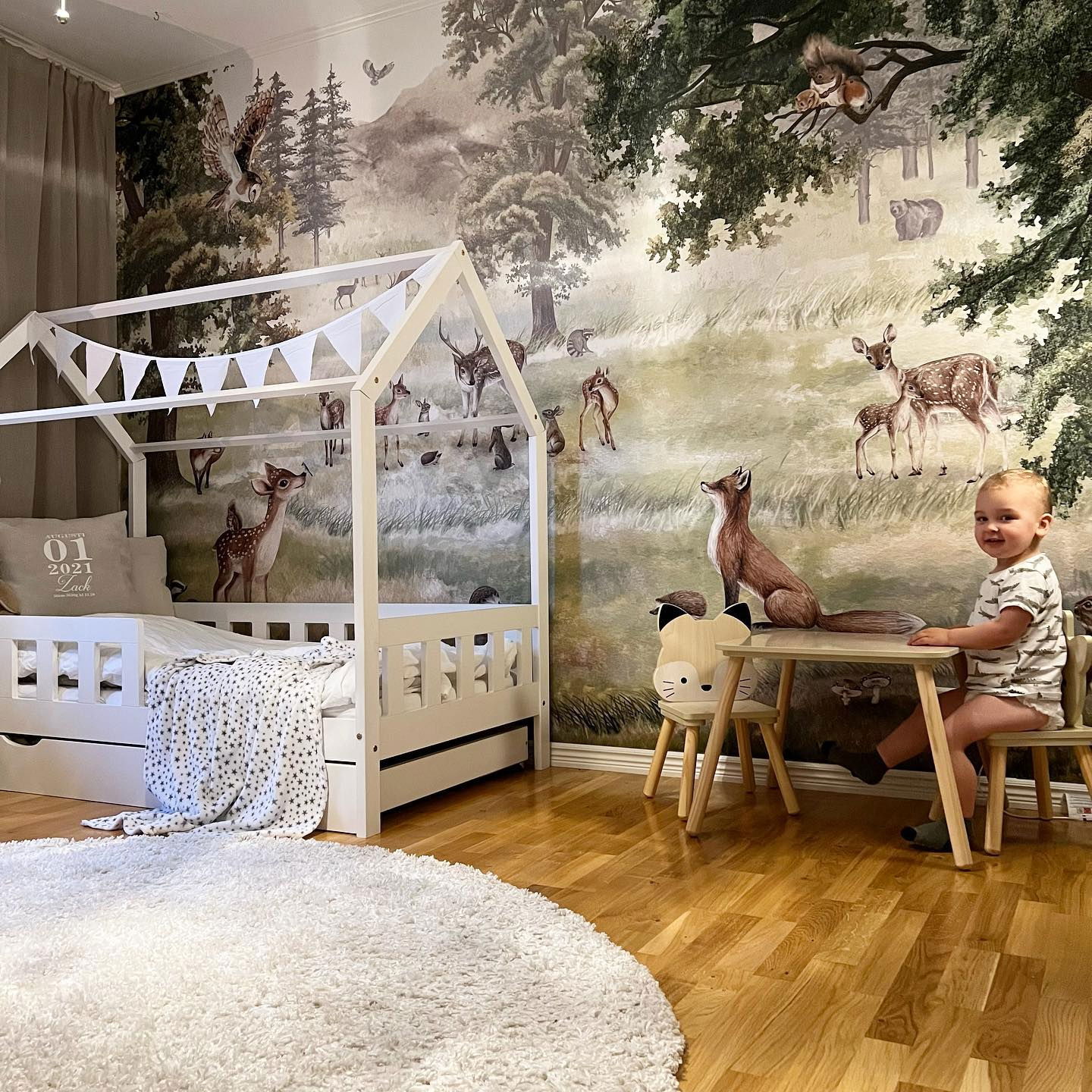 Chambre d'enfant avec une peinture murale sur le thème de la forêt, un lit cabane blanc et une petite table avec une chaise renard.