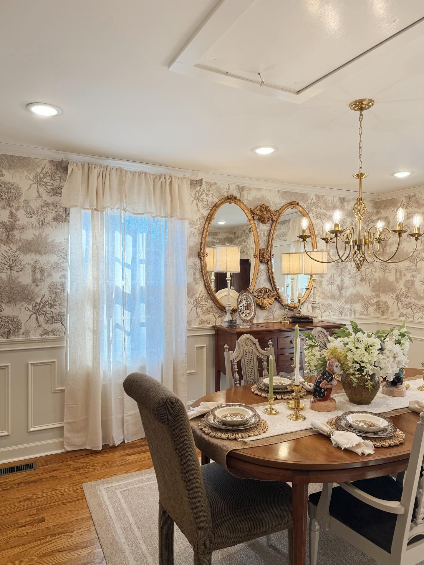 Dining room with a wooden table set with plates & candles. Brown toile wallpaper, gold oval mirrors, a brass chandelier.