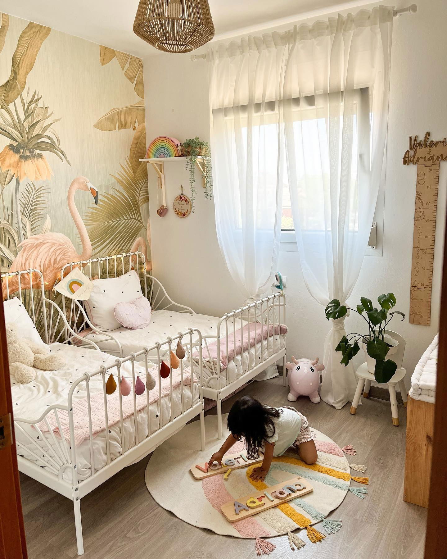 Niño juega con un rompecabezas de madera con su nombre en una alfombra de arcoíris en un dormitorio con camas individuales y papel tapiz de flamencos.
