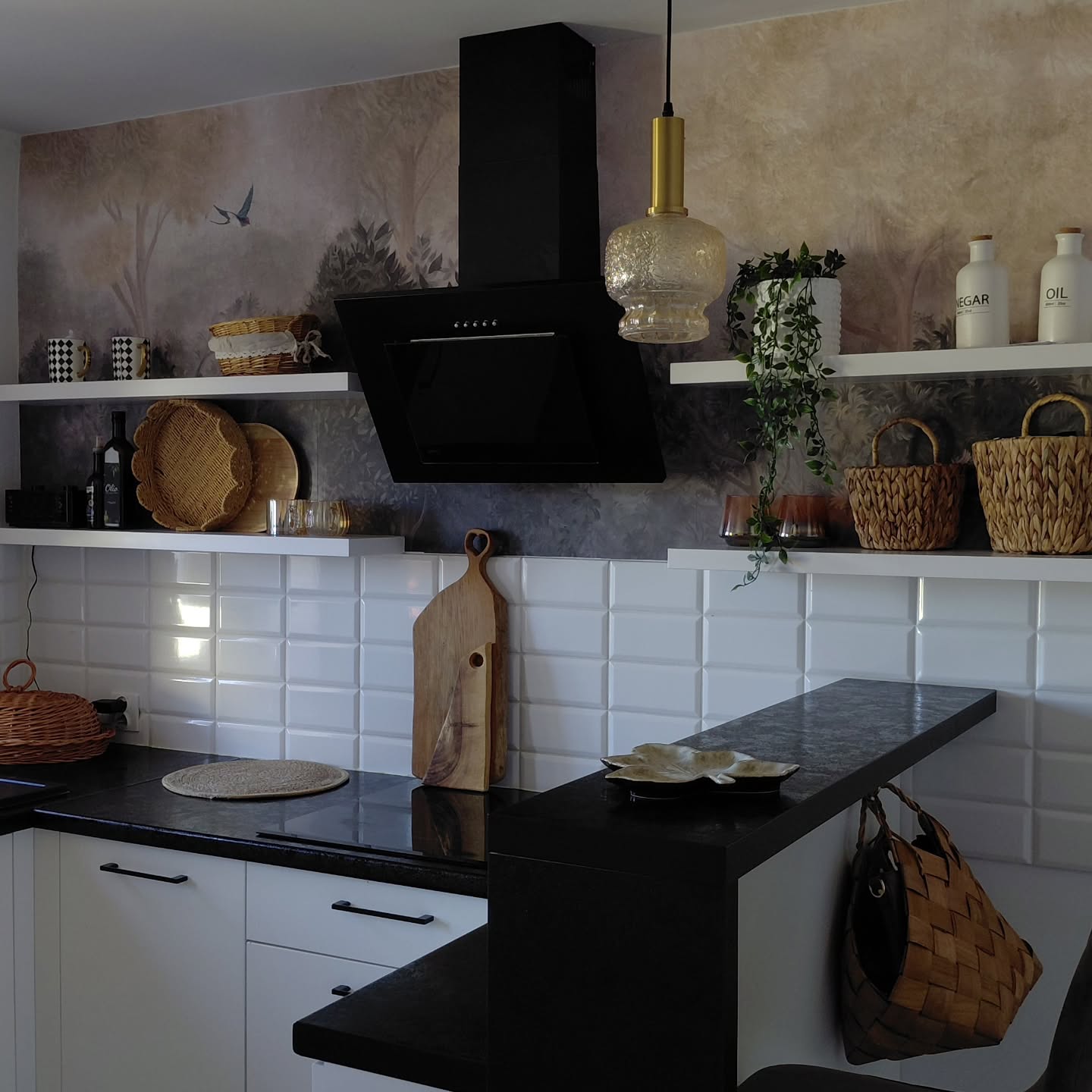 Una cucina moderna con una cappa aspirante nera e una lampada a sospensione dorata appesa sopra. Piastrelle bianche ricoprono il paraschizzi e un piano di lavoro nero si trova sotto i pensili bianchi. Sulla parete dietro la cappa è presente una carta da parati con una debole scena di foresta, con mensole bianche che contengono cesti e oggetti decorativi.