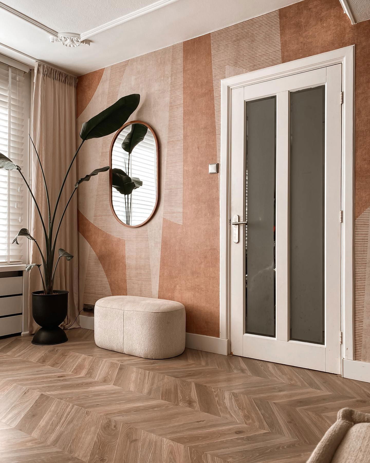 Interior with herringbone floor, patterned wallpaper, and white door. Potted plant and oval mirror accent the wall.