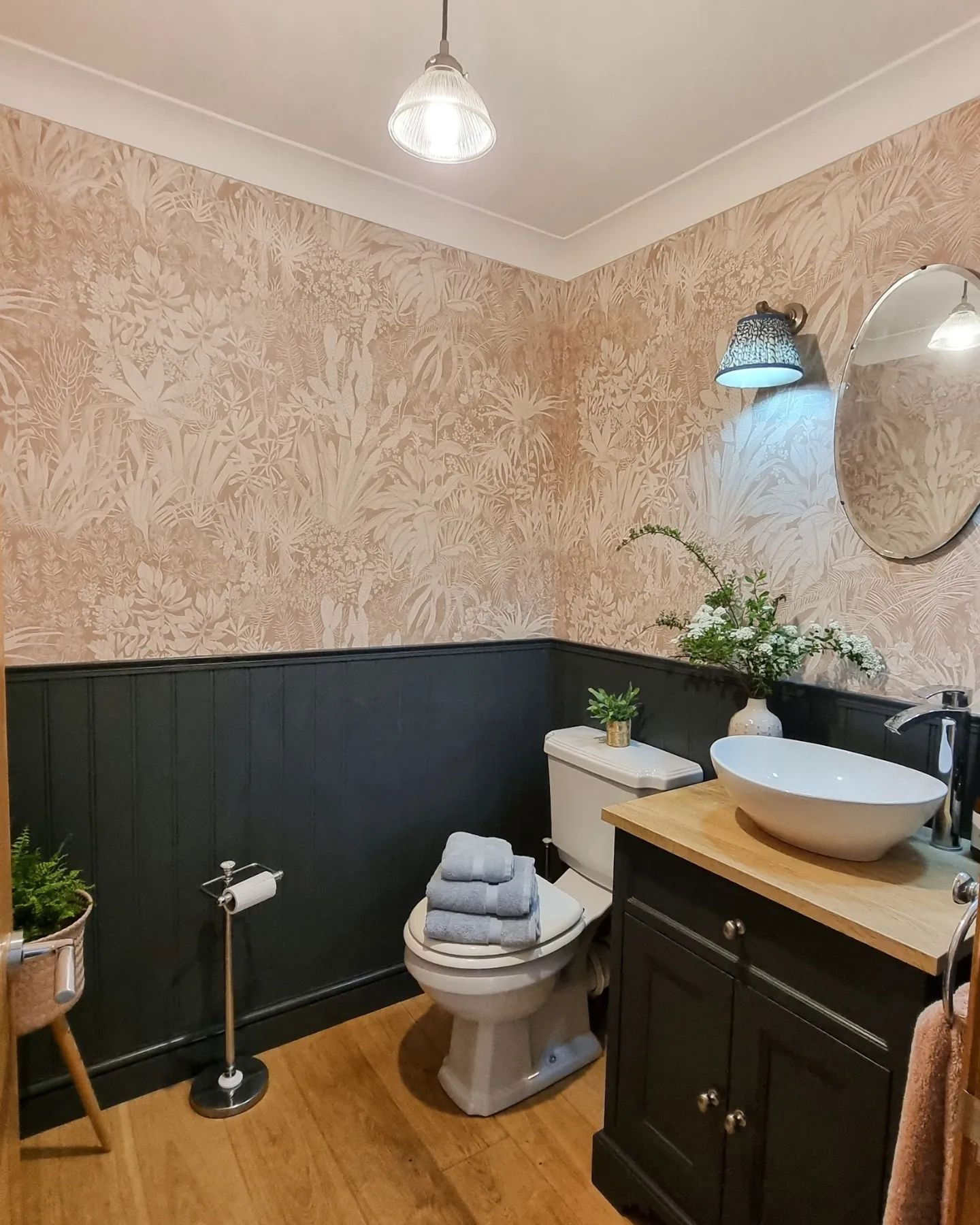 Baño pequeño con papel tapiz floral, revestimiento de madera negro y pisos de madera. Inodoro con toallas apiladas, tocador con lavabo tipo vasija.