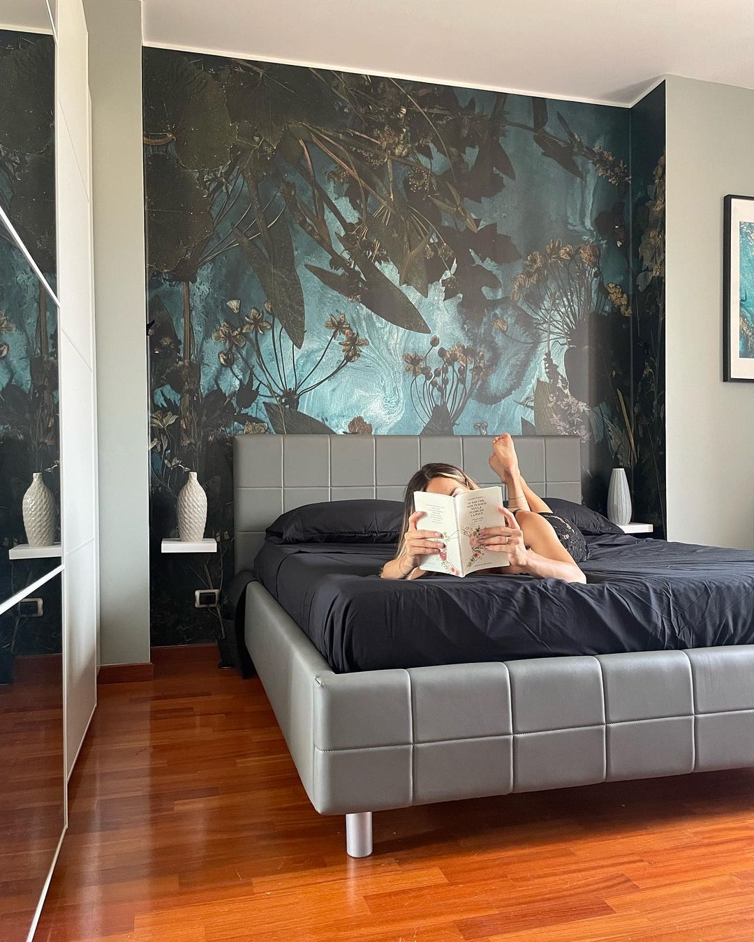 Woman reading on a gray upholstered bed with black sheets and a floral accent wall.