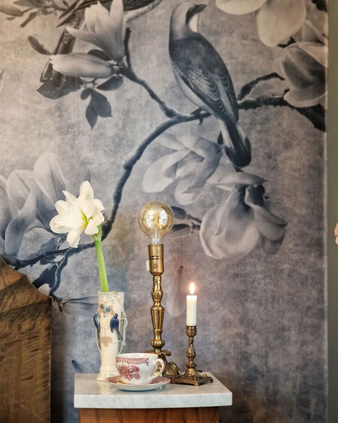 Stillleben mit Blume, Teetasse und Kerzen auf einem Marmortisch vor einer floralen Vogel-Tapete.