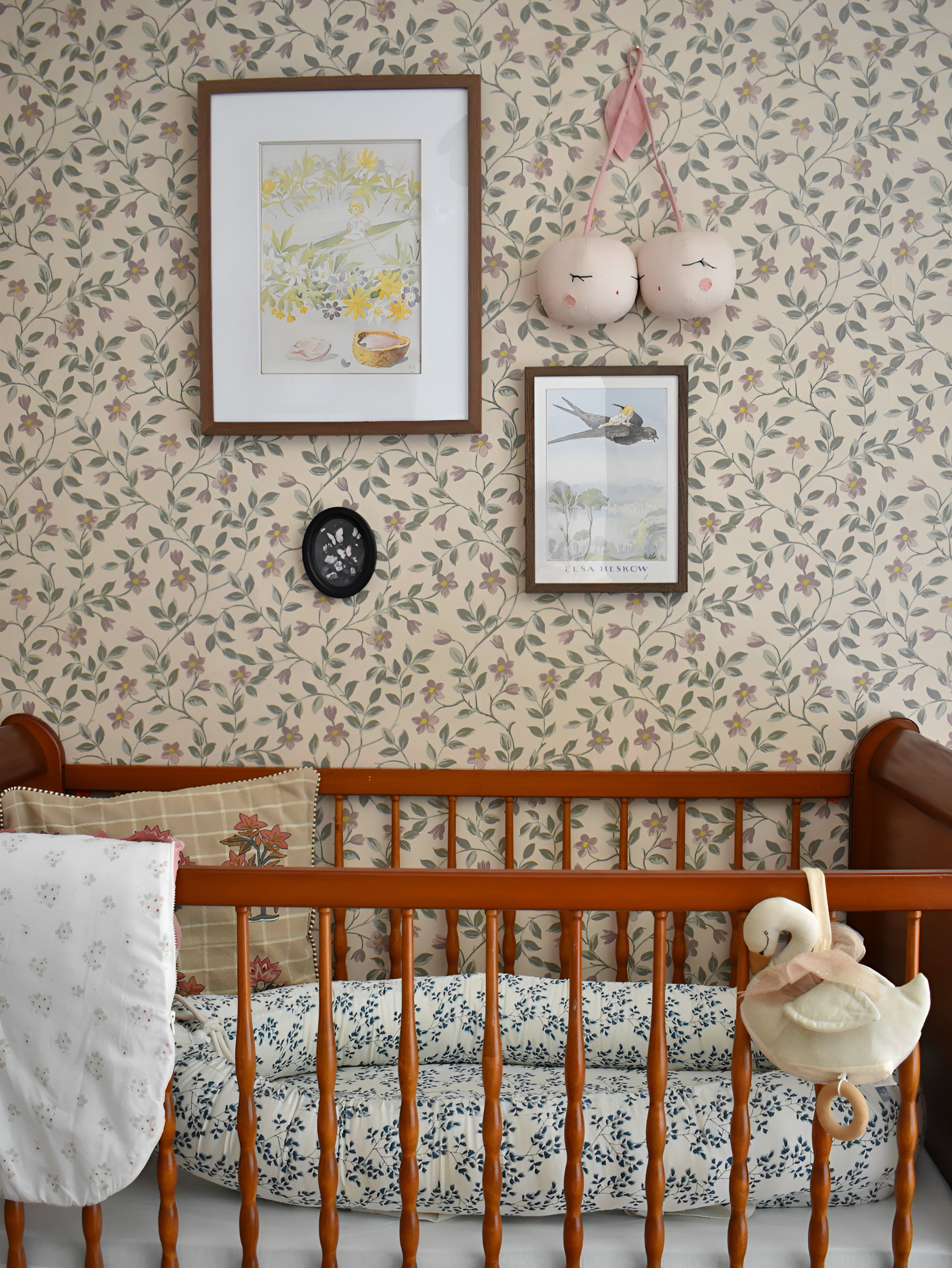 A wooden crib with blue floral bedding and a white swan toy. Cream floral wallpaper with various wall decorations.