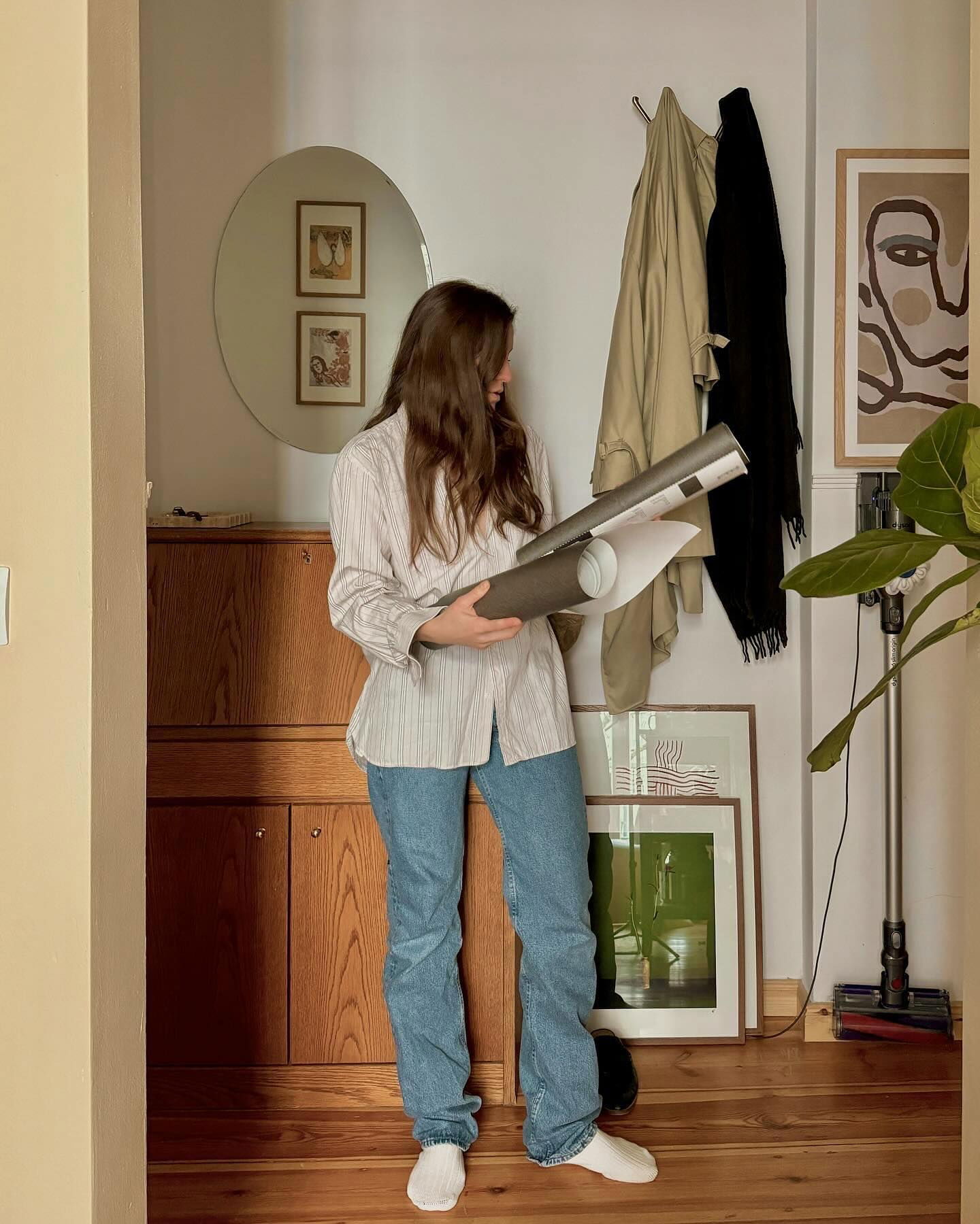 Eine Frau hält gerollte Poster in einem Flur mit Mänteln, Kunst und einem Staubsauger.