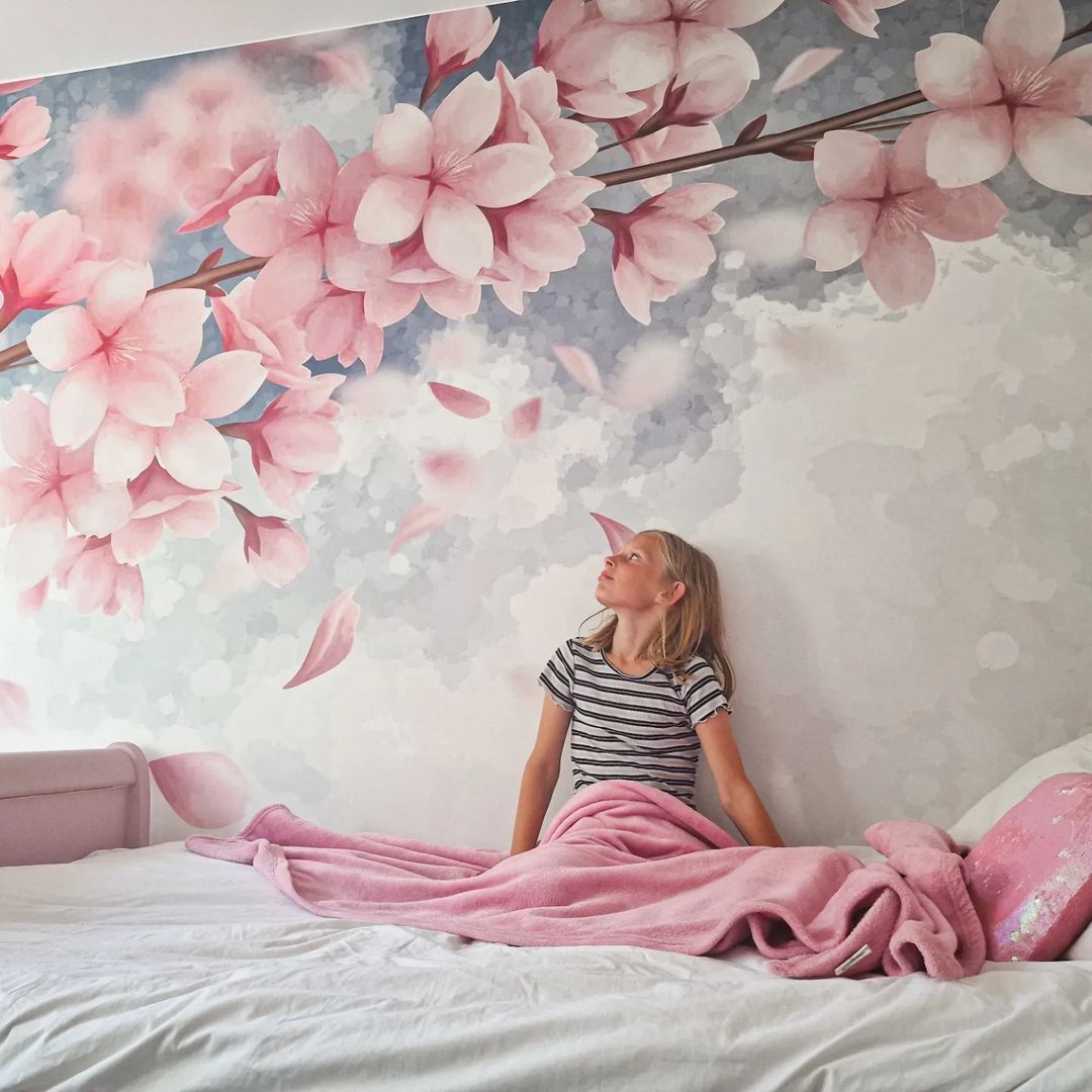 Girl in bed looks at cherry blossom wall mural. Pink blanket and pillow on white sheets.
