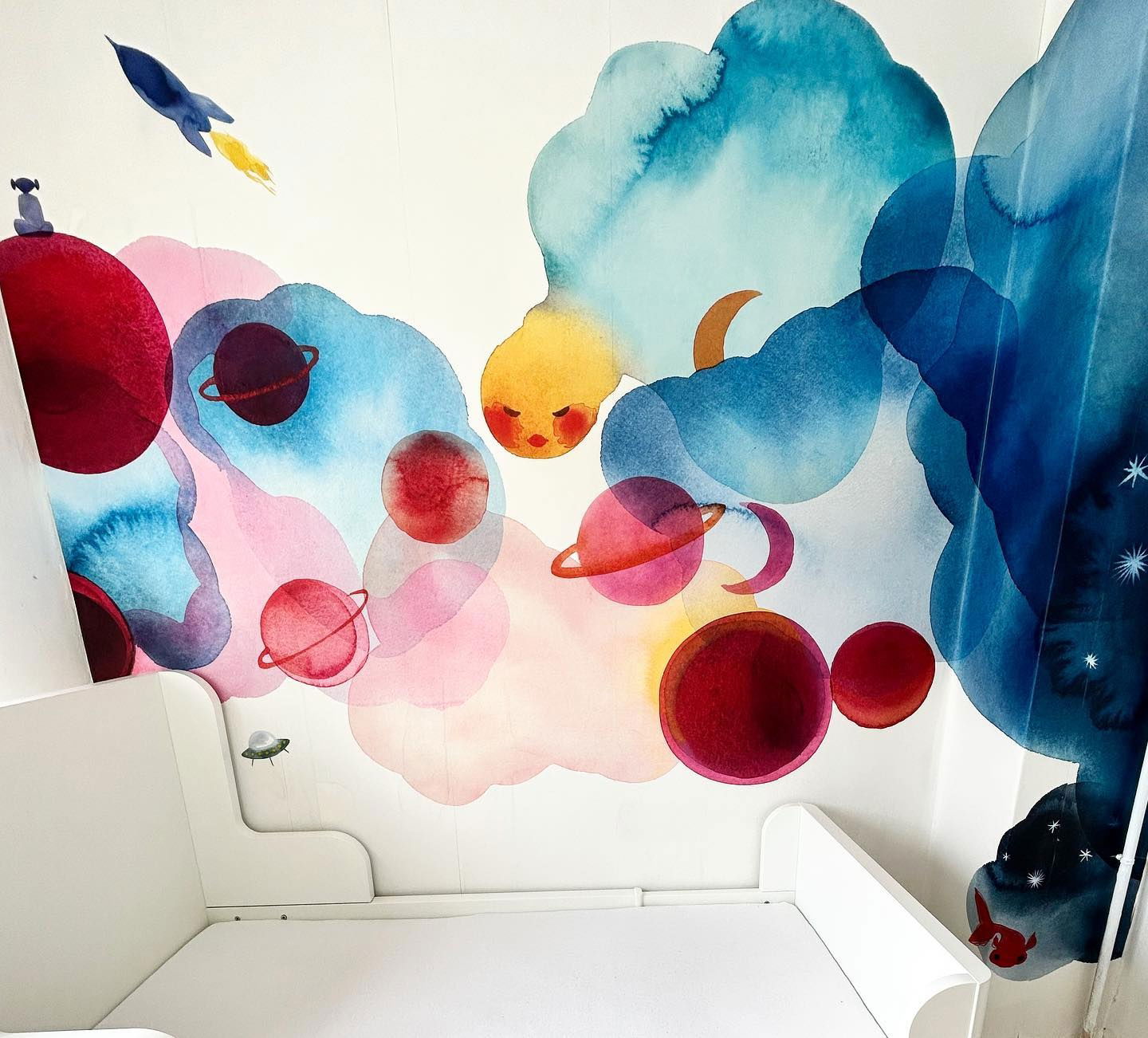 Nursery with white crib and watercolor space-themed wall art featuring planets, clouds, and a rocket.