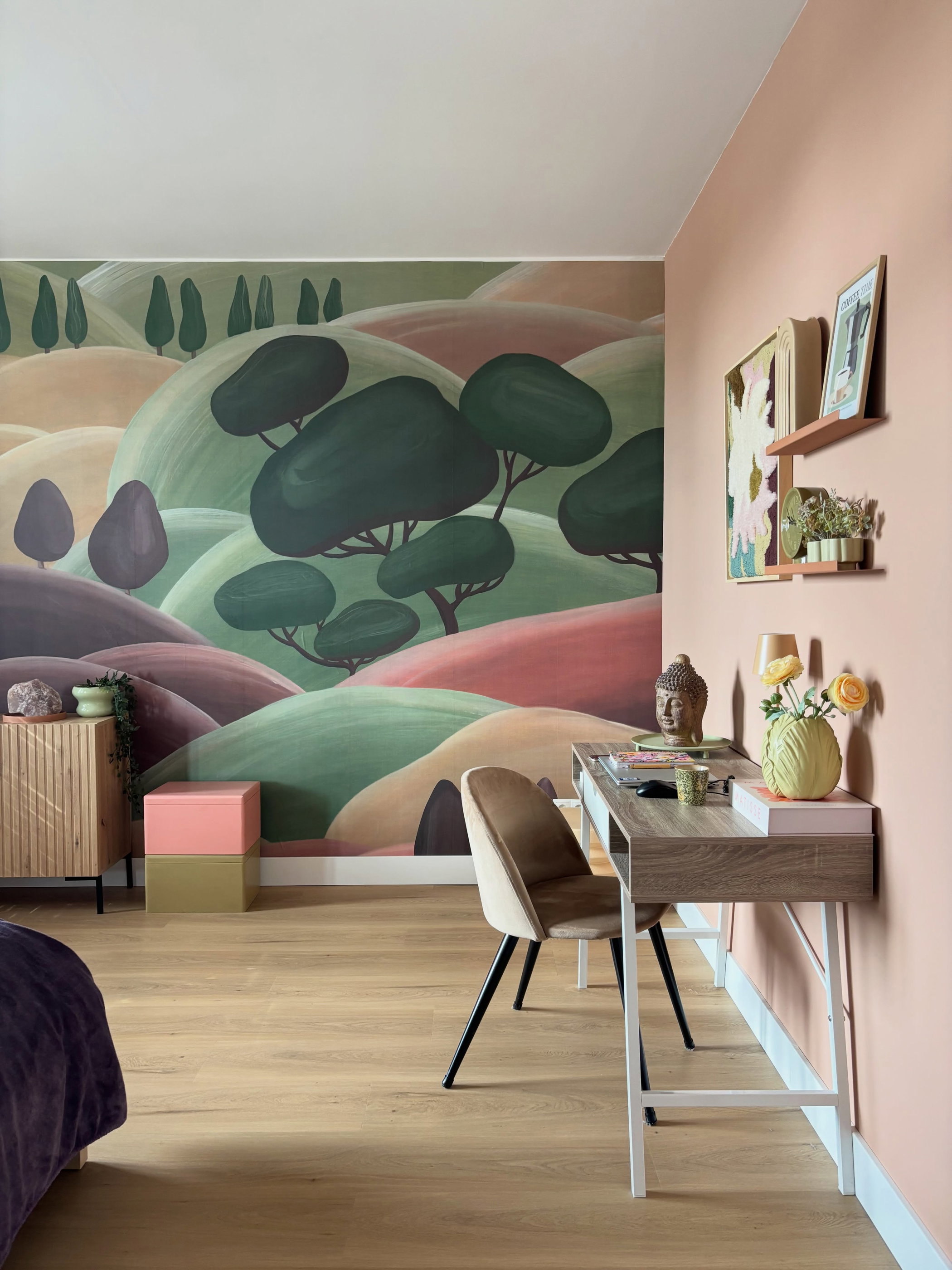 A room with a mural of green and pink rolling hills and stylized trees. A light wood desk and beige chair against a peach wall, with a Buddha, yellow flowers, and shelves.