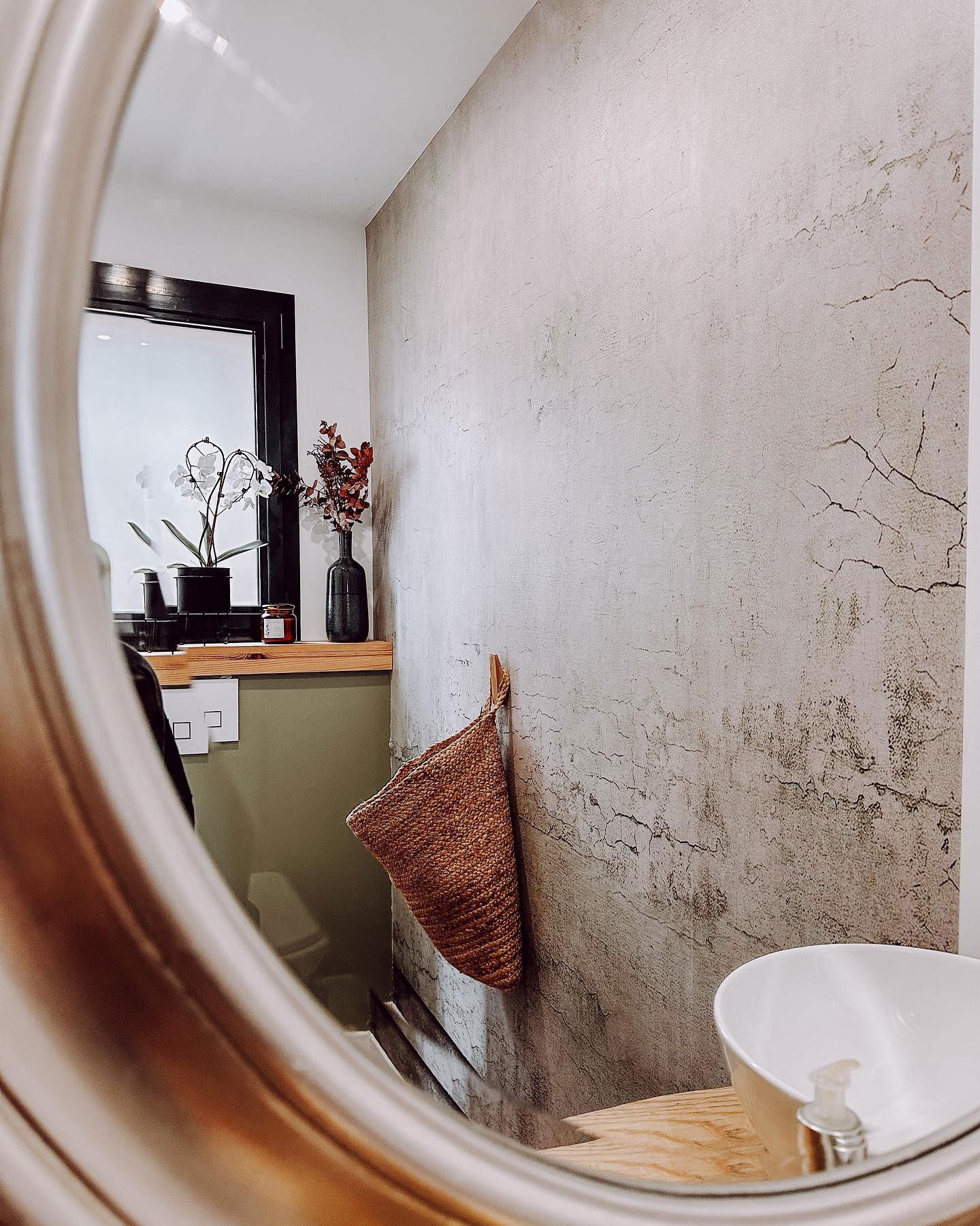 Bathroom interior reflected in mirror. Woven basket hangs on textured wall near a white sink. Window with plants in background.